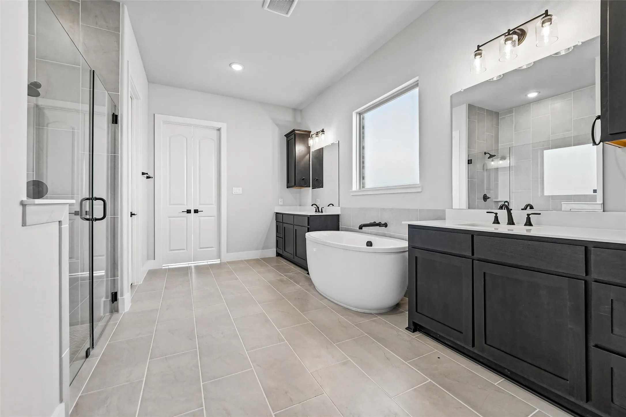 Dual vanity with white countertops, freestanding soaking tub, frameless glass shower enclosure, and tiled flooring