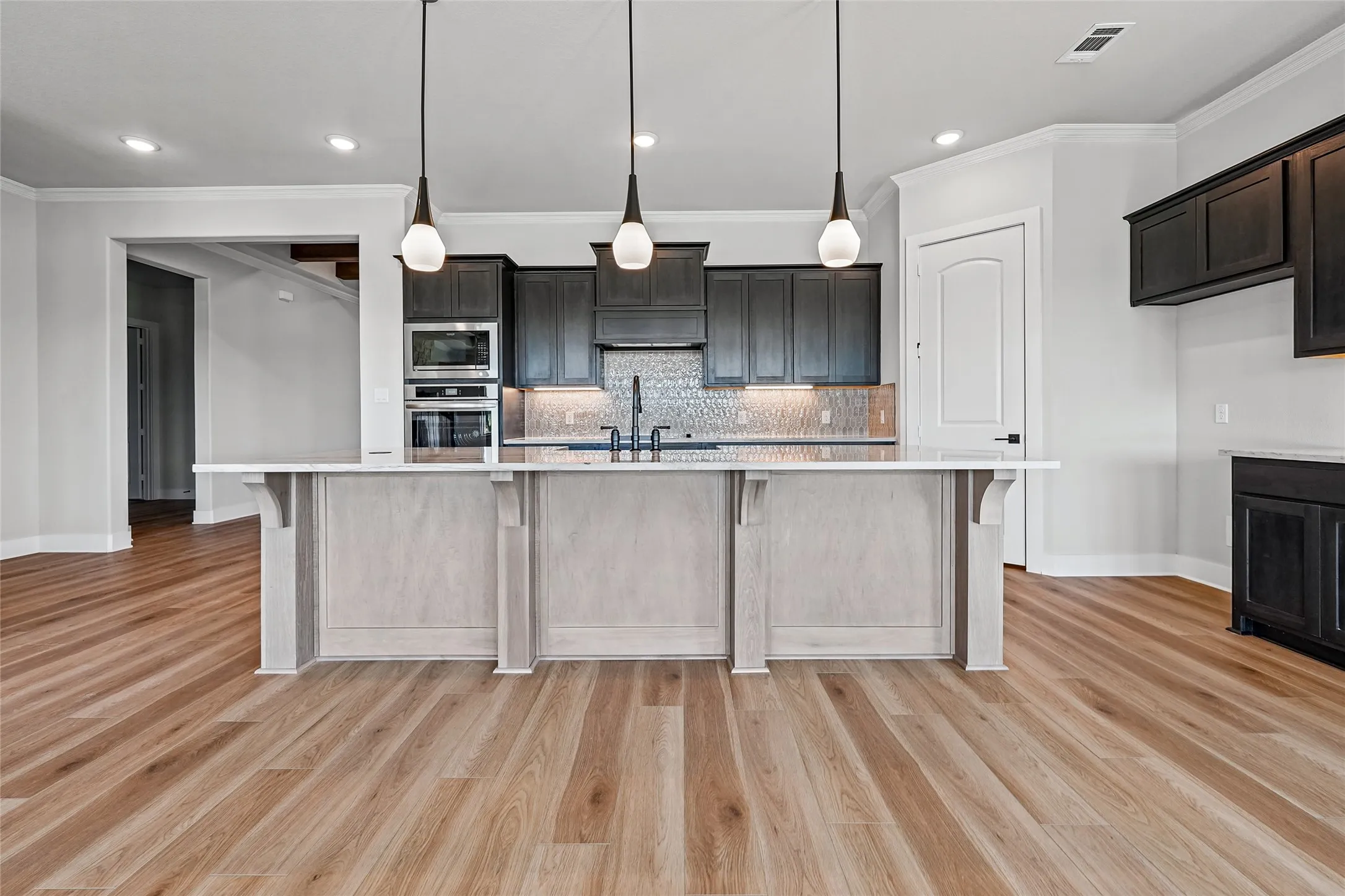 Expansive wood-finish flooring, large kitchen island with white countertop, dark wood cabinetry, built-in stainless steel appliances, and patterned tile backsplash