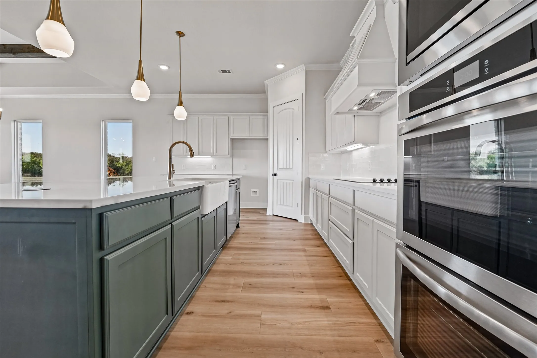 Spacious kitchen featuring a large central island with a farmhouse sink, white perimeter cabinetry, wood-finish flooring, and integrated stainless steel double ovens