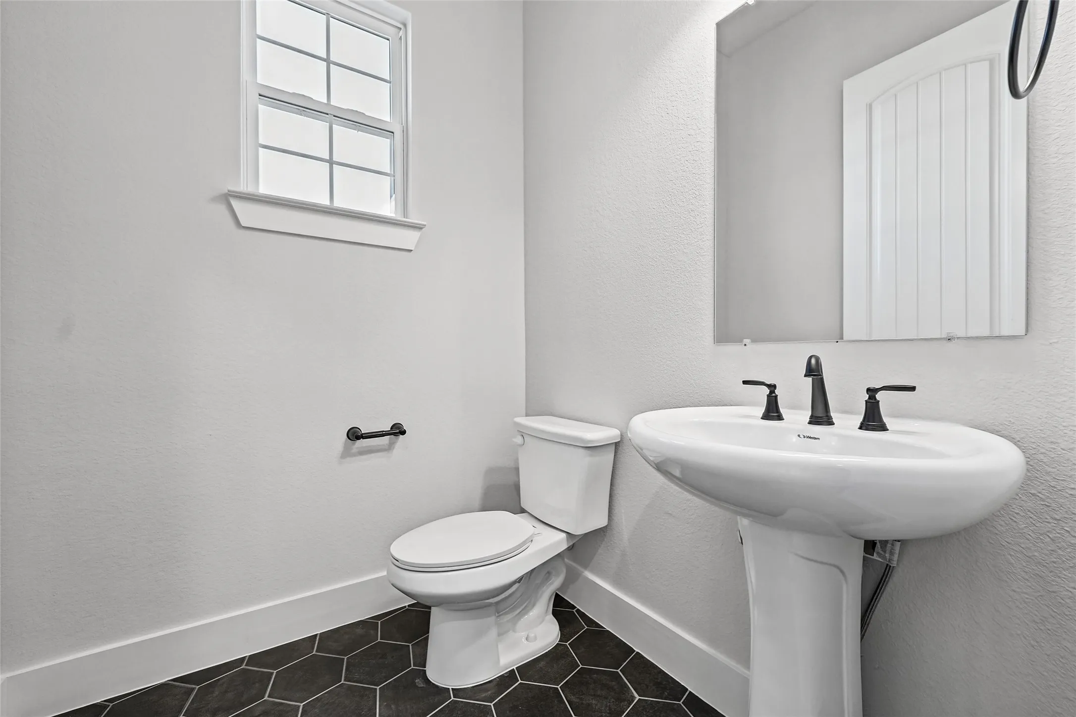 Hexagonal floor tiling, pedestal sink, wall-mounted mirror, matte black fixtures, and a white framed window