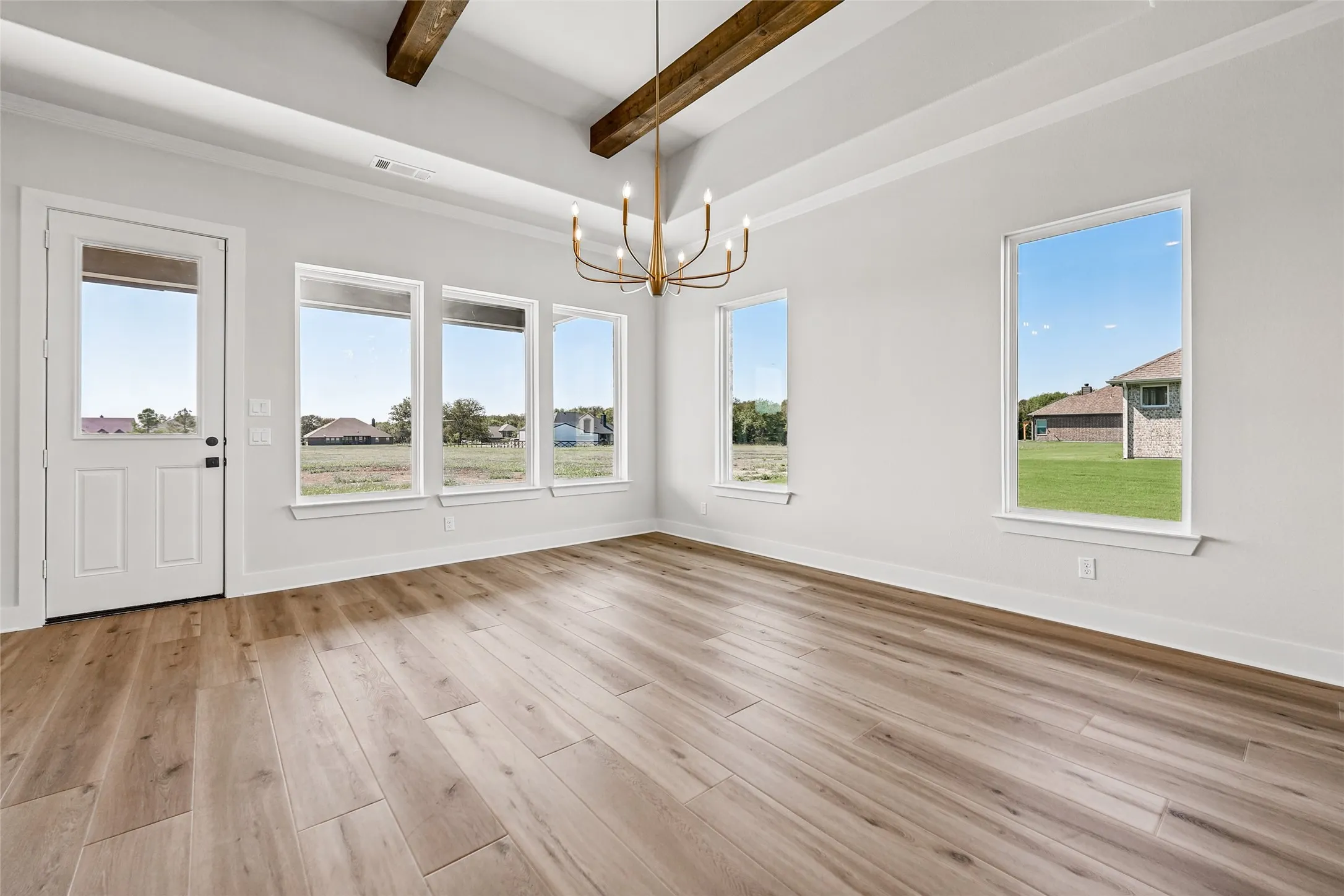 Vaulted ceiling with exposed wood beams, wood-finish flooring, abundant natural light, brushed gold chandelier, and a white paneled door with a glass insert