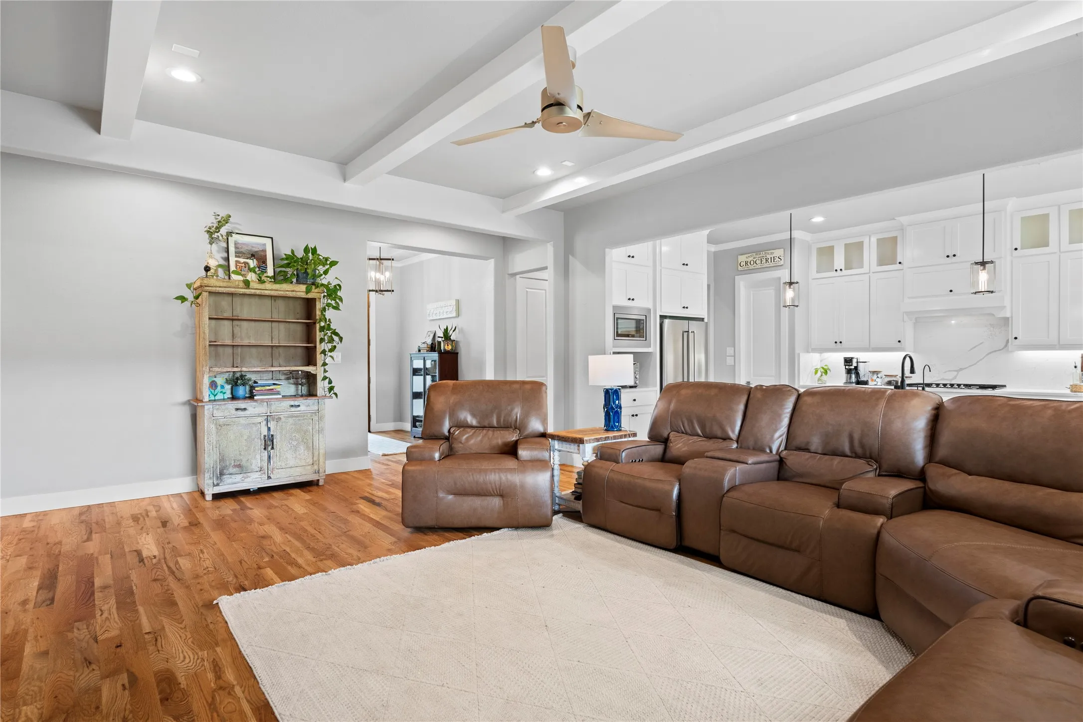 Open-concept living space featuring wood-finish flooring, a tray ceiling with recessed lighting and a ceiling fan, and white cabinetry in the background kitchen