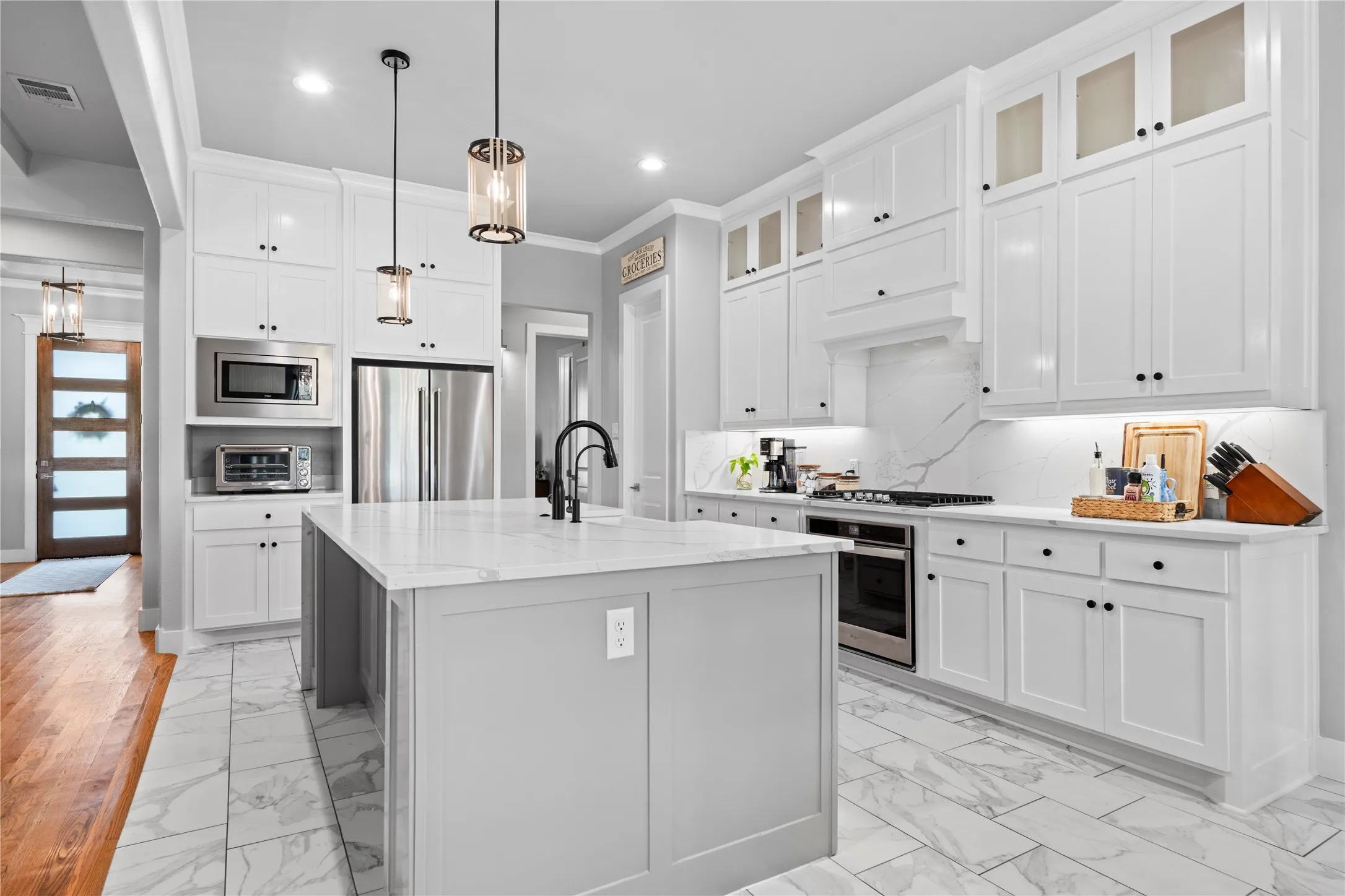 Modern kitchen featuring a central island with a waterfall-edge countertop, stainless steel appliances, white shaker cabinetry, and marble-look tile flooring