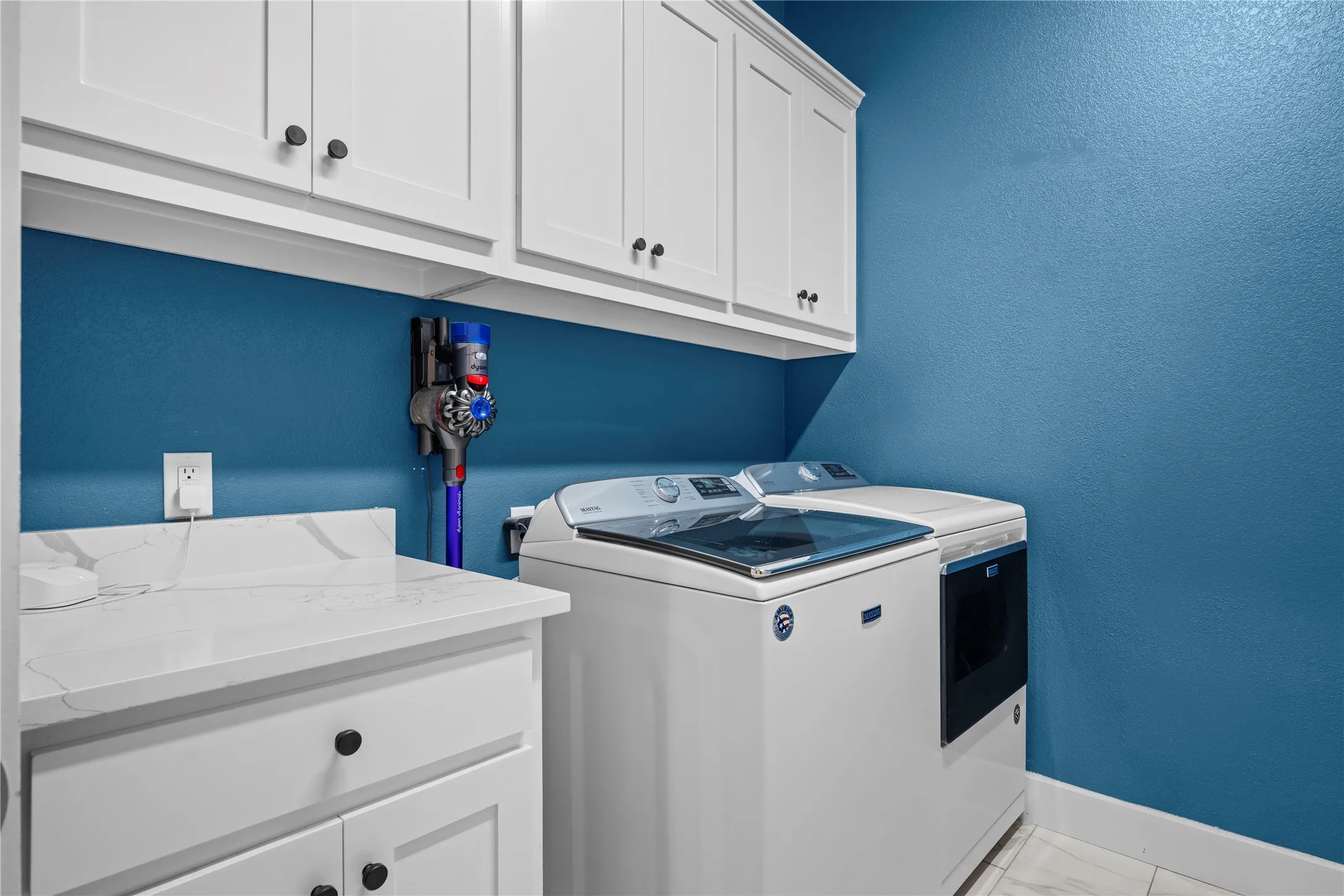 Laundry area featuring white cabinetry with black hardware, a light-colored countertop with veining, and a vibrant blue wall color