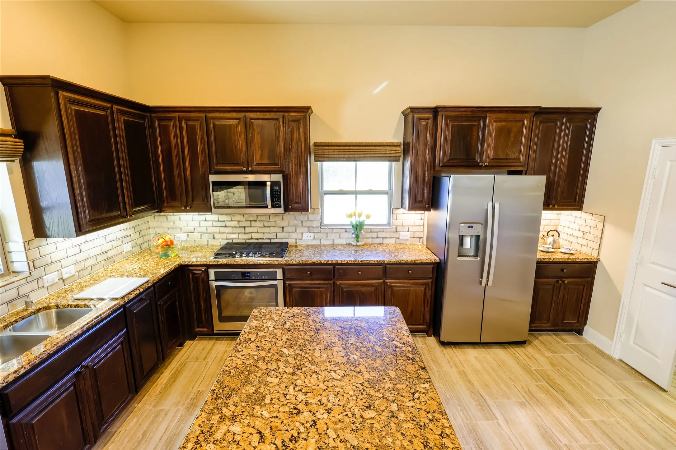 Kitchen featuring an expansive island with granite-finish countertop, dark wood-finish cabinetry, stainless steel appliances, and a subway tile backsplash
