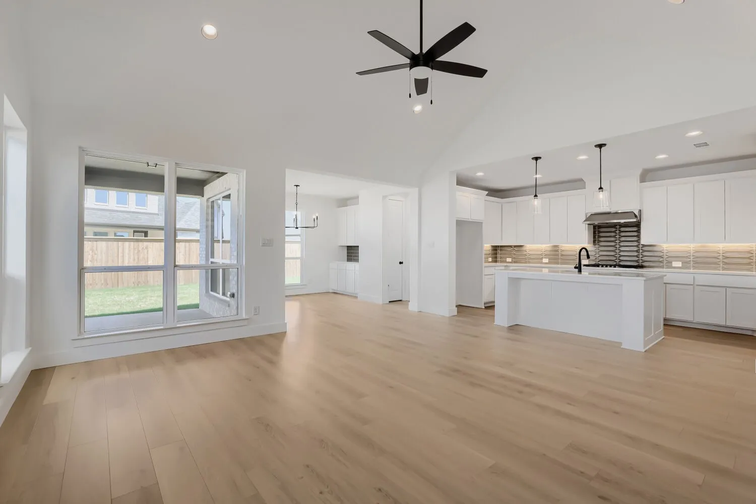 Vaulted ceiling with a ceiling fan, light wood-finish flooring, white kitchen cabinetry, kitchen island with integrated sink, and an outdoor patio with a fenced yard