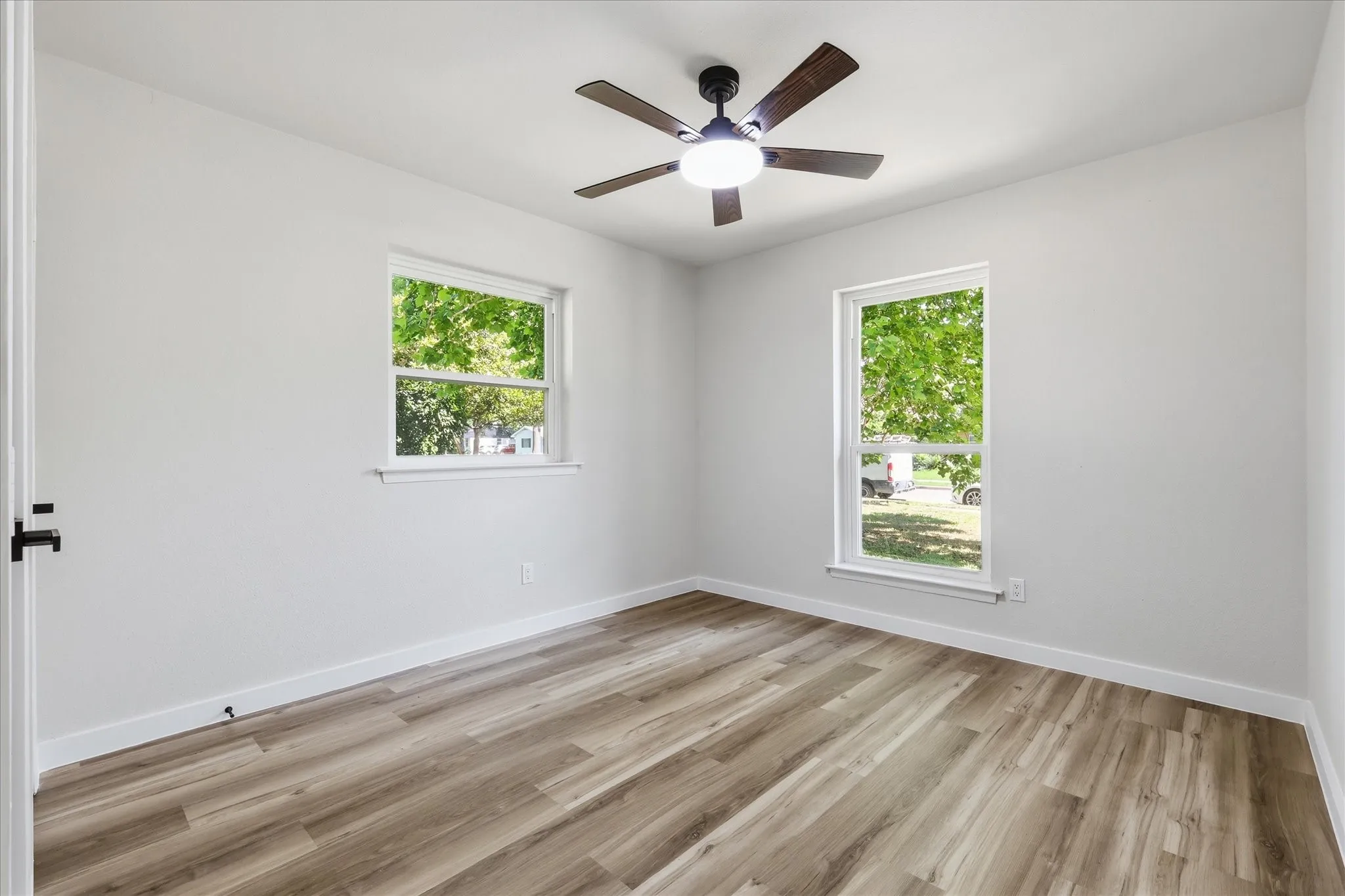 Bright room featuring wood-finish flooring, white walls, two window openings, a ceiling fan with integrated lighting, and black hardware