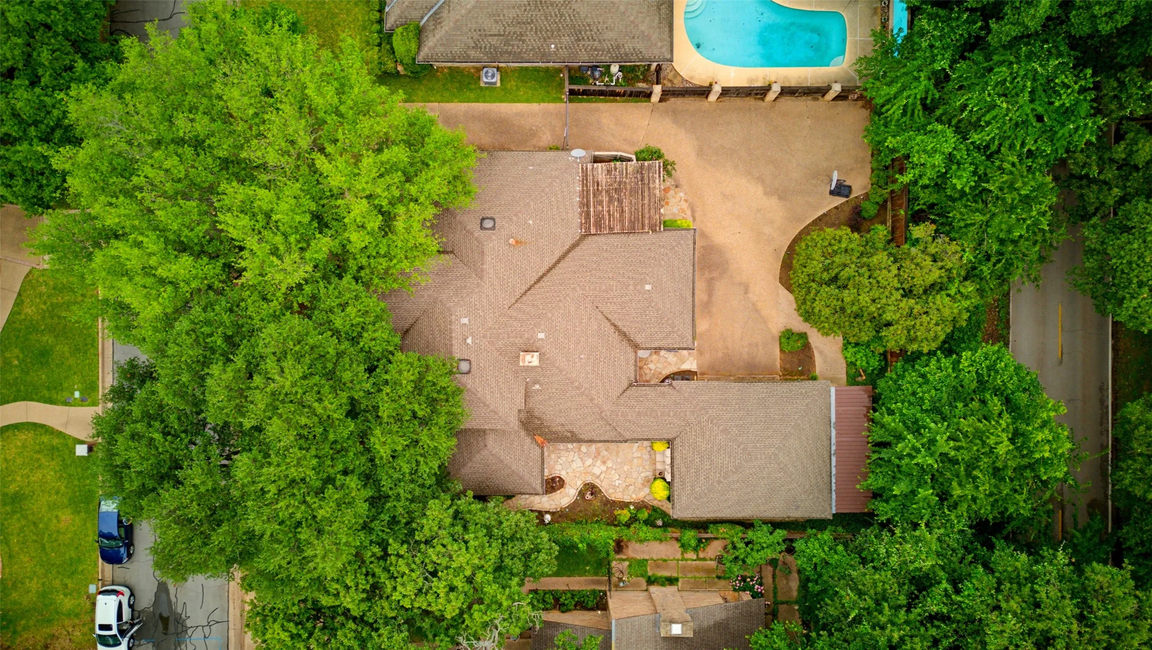 Overhead property perspective showcasing a substantial residential structure with a brown shingle roof, a generously sized driveway, and mature trees providing ample foliage