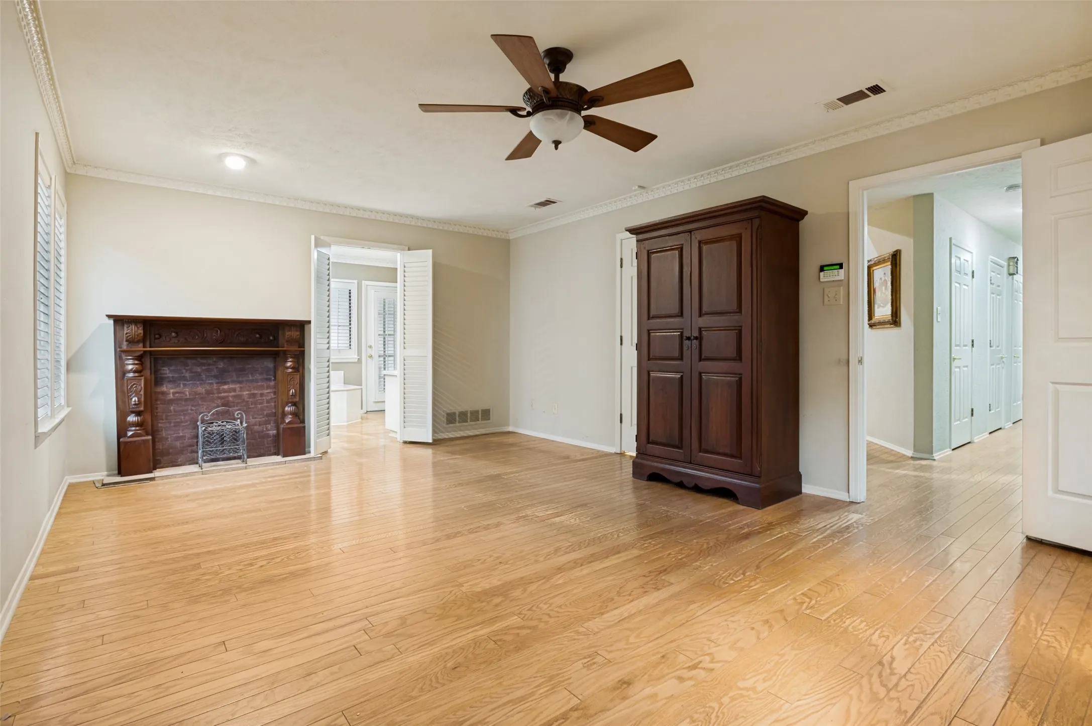 Spacious room featuring wood-finish flooring, a decorative wood mantel over a brick fireplace, crown molding, and a ceiling fan with integrated lighting
