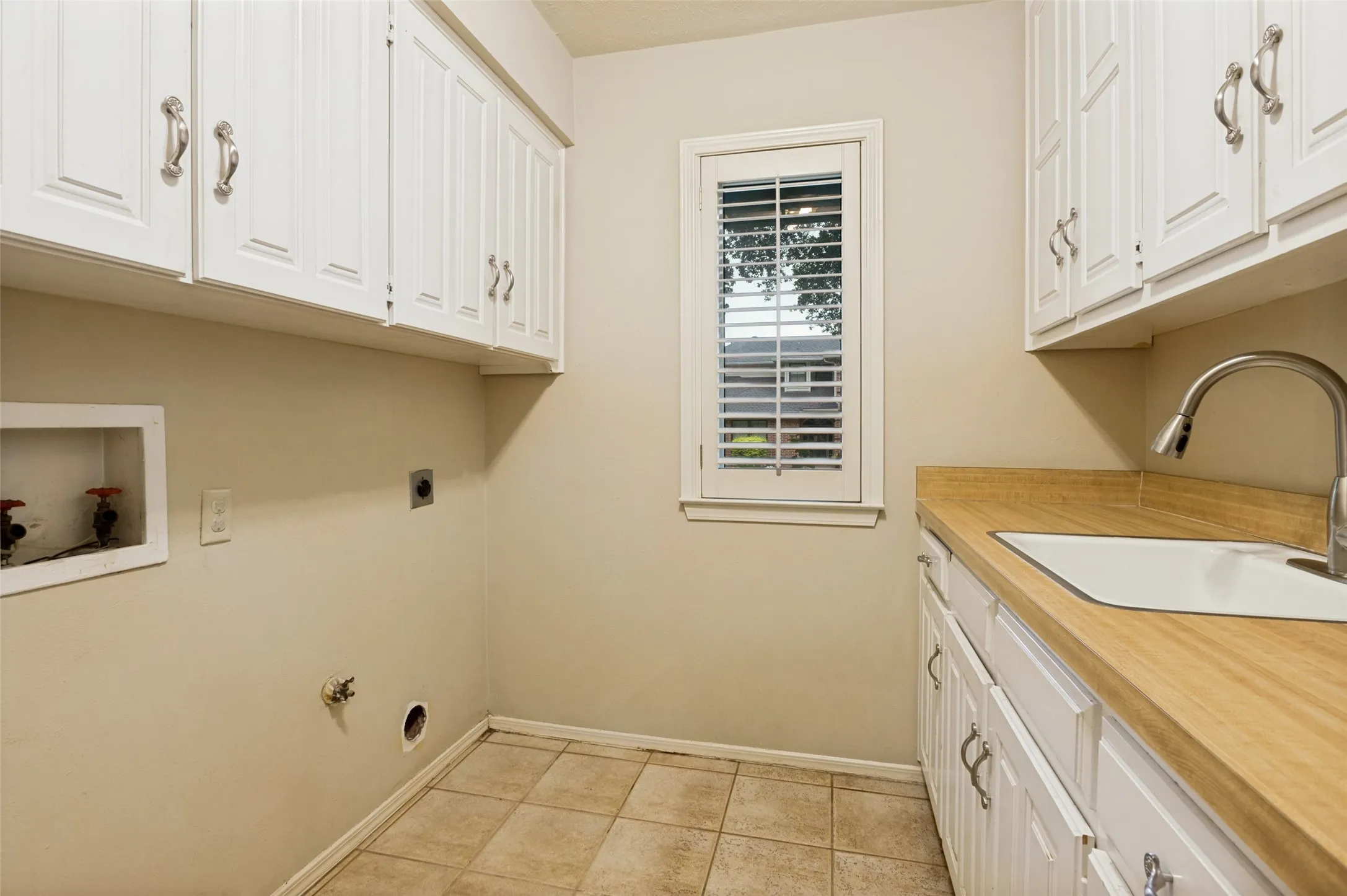 Laundry room featuring a utility sink, wood-finish countertop, white cabinetry with silver-tone hardware, and a window with plantation shutters