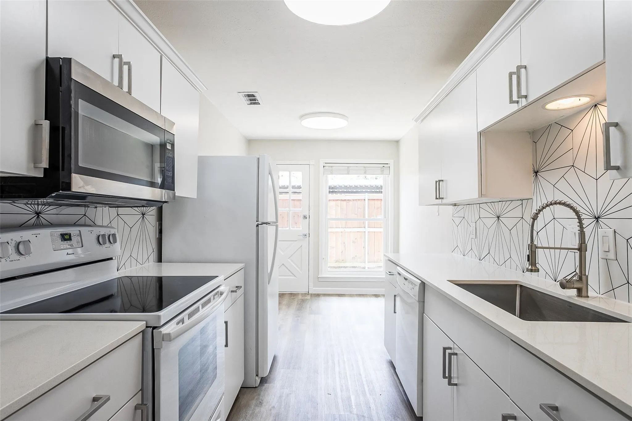 Galley kitchen featuring white cabinetry with brushed nickel hardware, stainless steel appliances, geometric tile backsplash, light countertops, and wood-finish flooring
