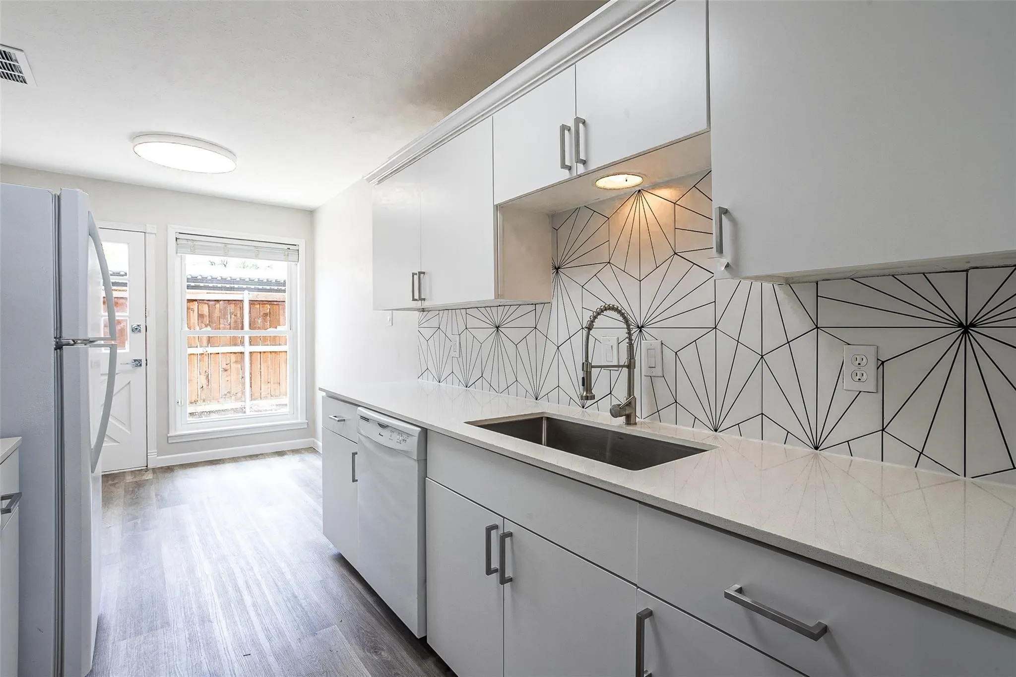 Modern kitchen featuring white cabinetry, light-toned countertops, a geometric pattern backsplash, wood-finish flooring, and a stainless steel sink with a gooseneck faucet