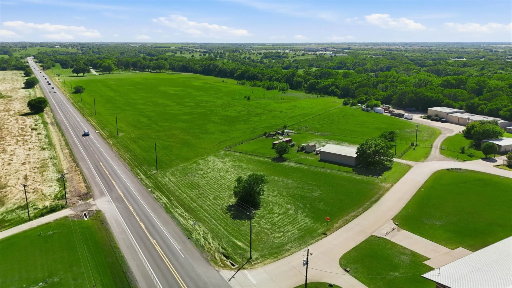Expansive green acreage featuring a two-lane paved road, mature trees, and several accessory structures