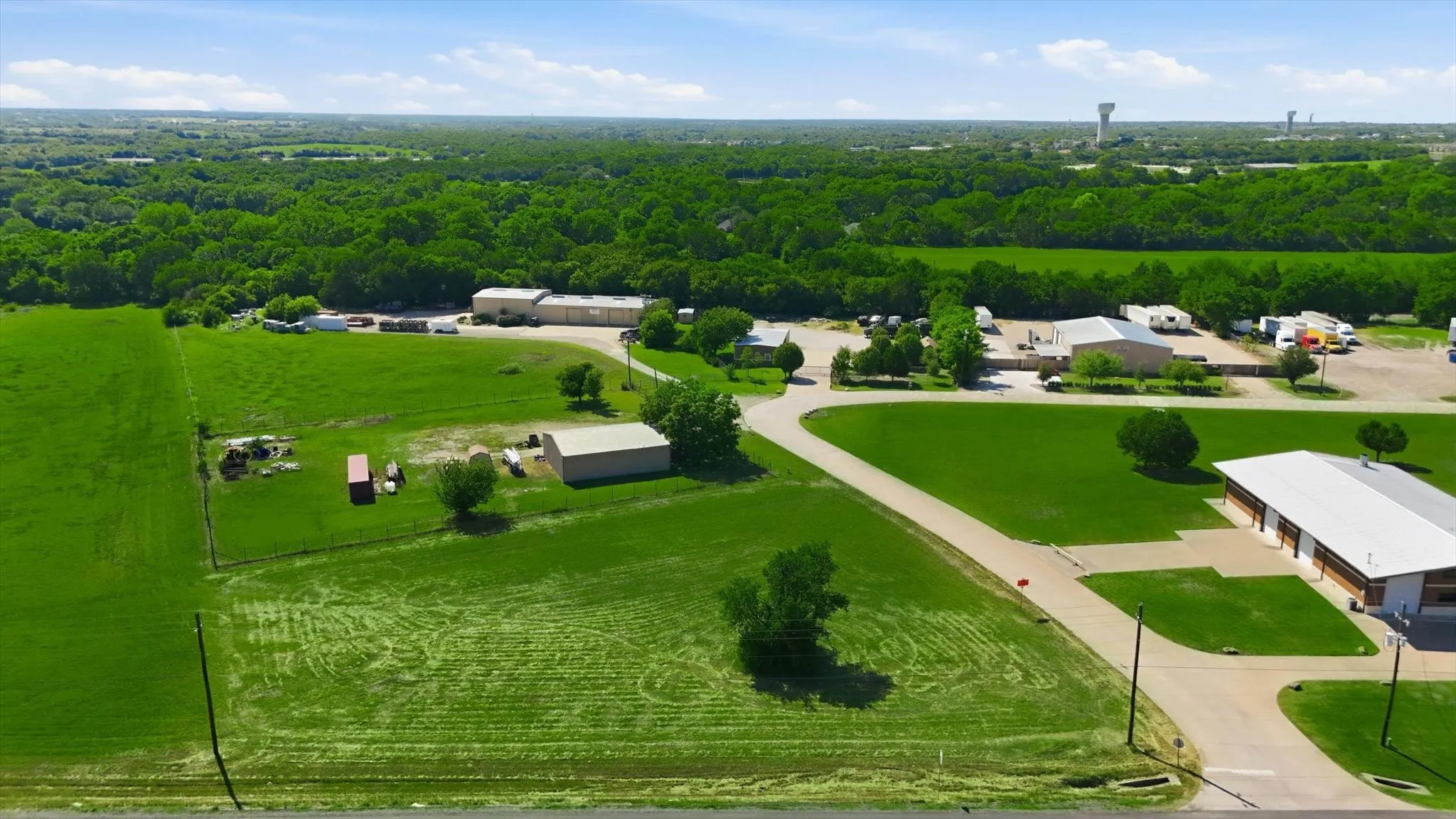 Expansive green acreage featuring multiple commercial-style buildings with light-colored roofs, paved driveways, and surrounding natural tree lines