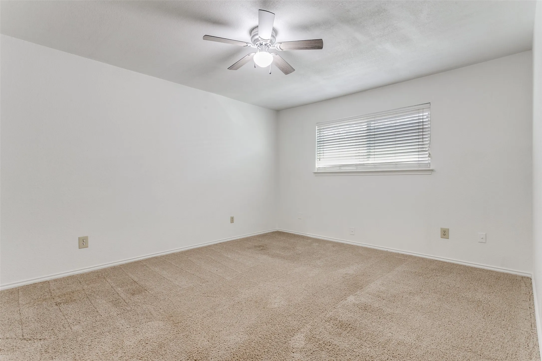 Spacious room featuring neutral carpeting, a large window with horizontal blinds, a ceiling fan with integrated lighting, and light-toned walls