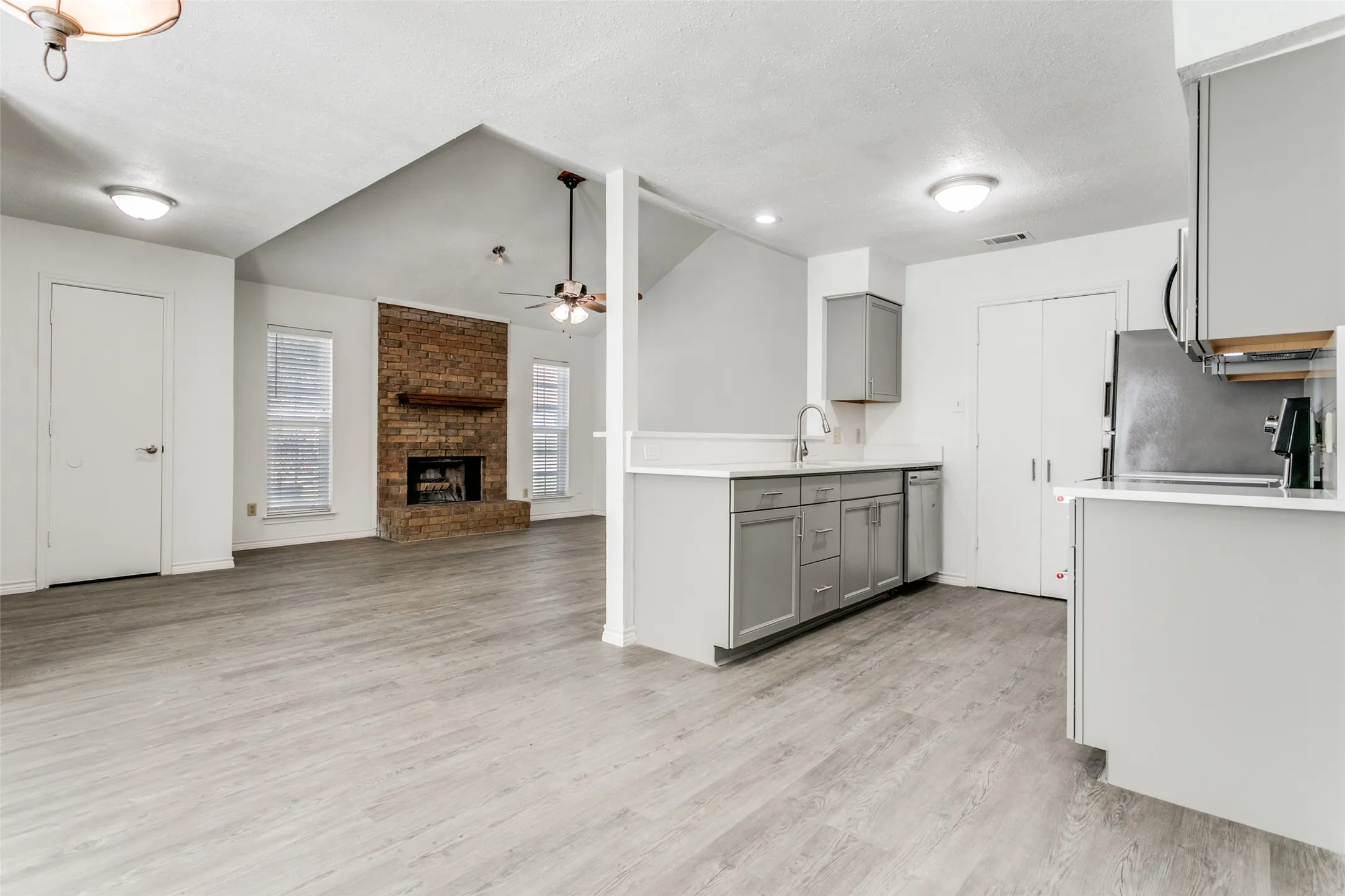 Open-concept living area featuring light wood-finish flooring throughout, a brick fireplace with a wood mantel, vaulted ceilings, and a ceiling fan