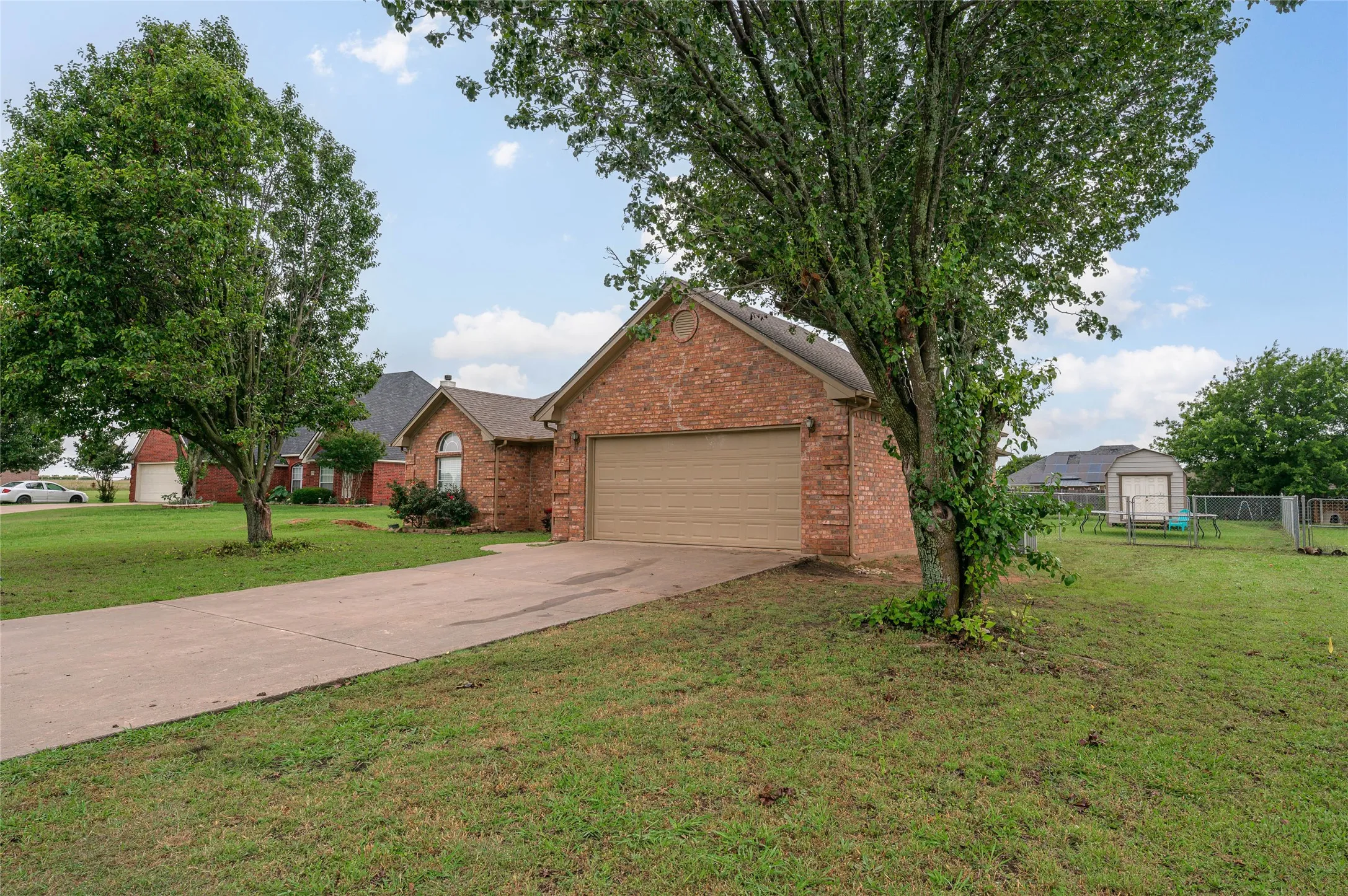 Brick exterior residence featuring an attached garage with a tan door, a concrete driveway, a front lawn with mature trees, and a backyard with a storage shed