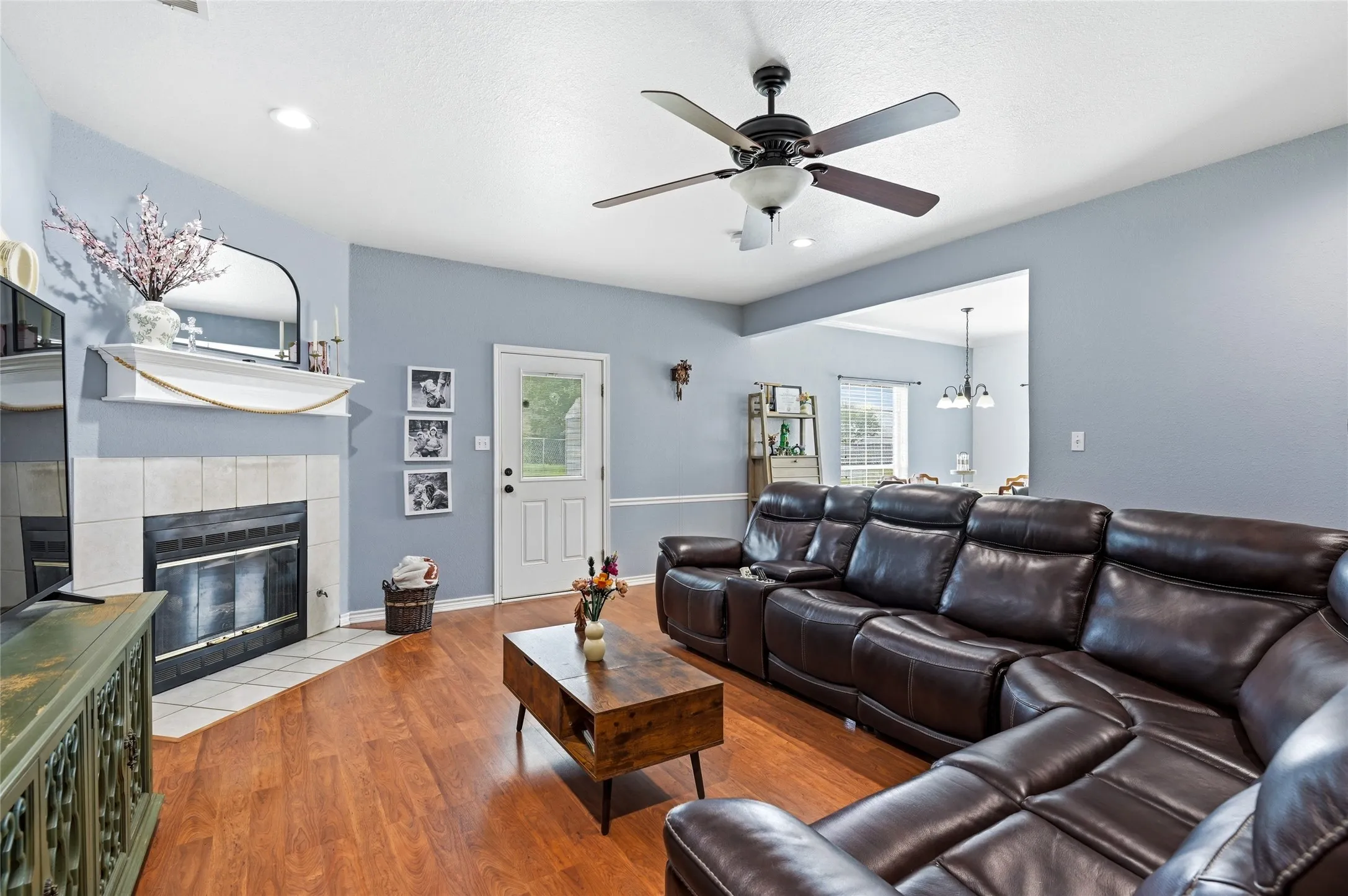 Living area featuring wood-finish flooring, a tiled fireplace with mantel, ceiling fan with integrated lighting, and a white paneled door with a glass insert