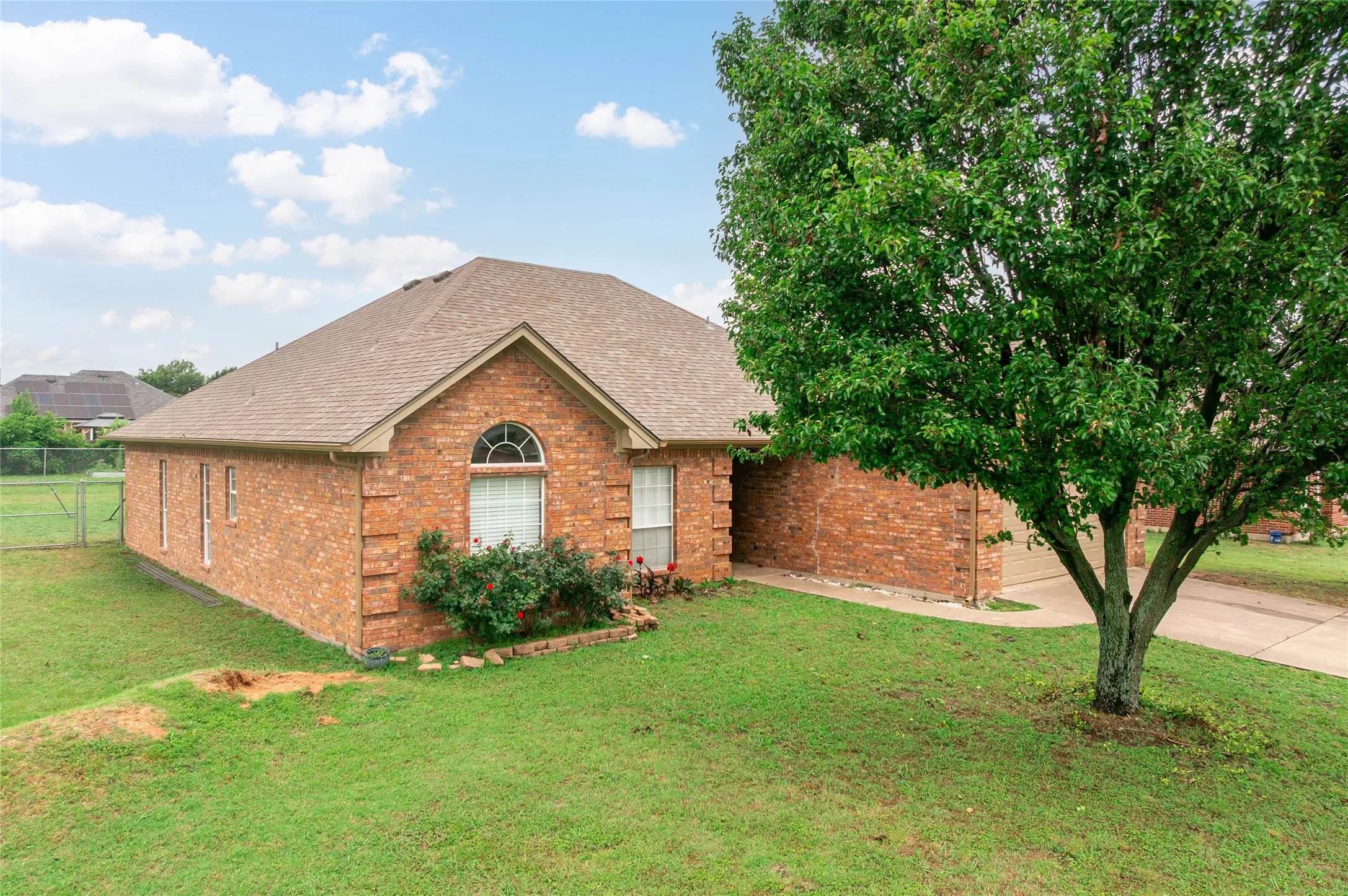 Brick exterior with an arched window, a brown shingle roof, established landscaping, and a chain-link fence