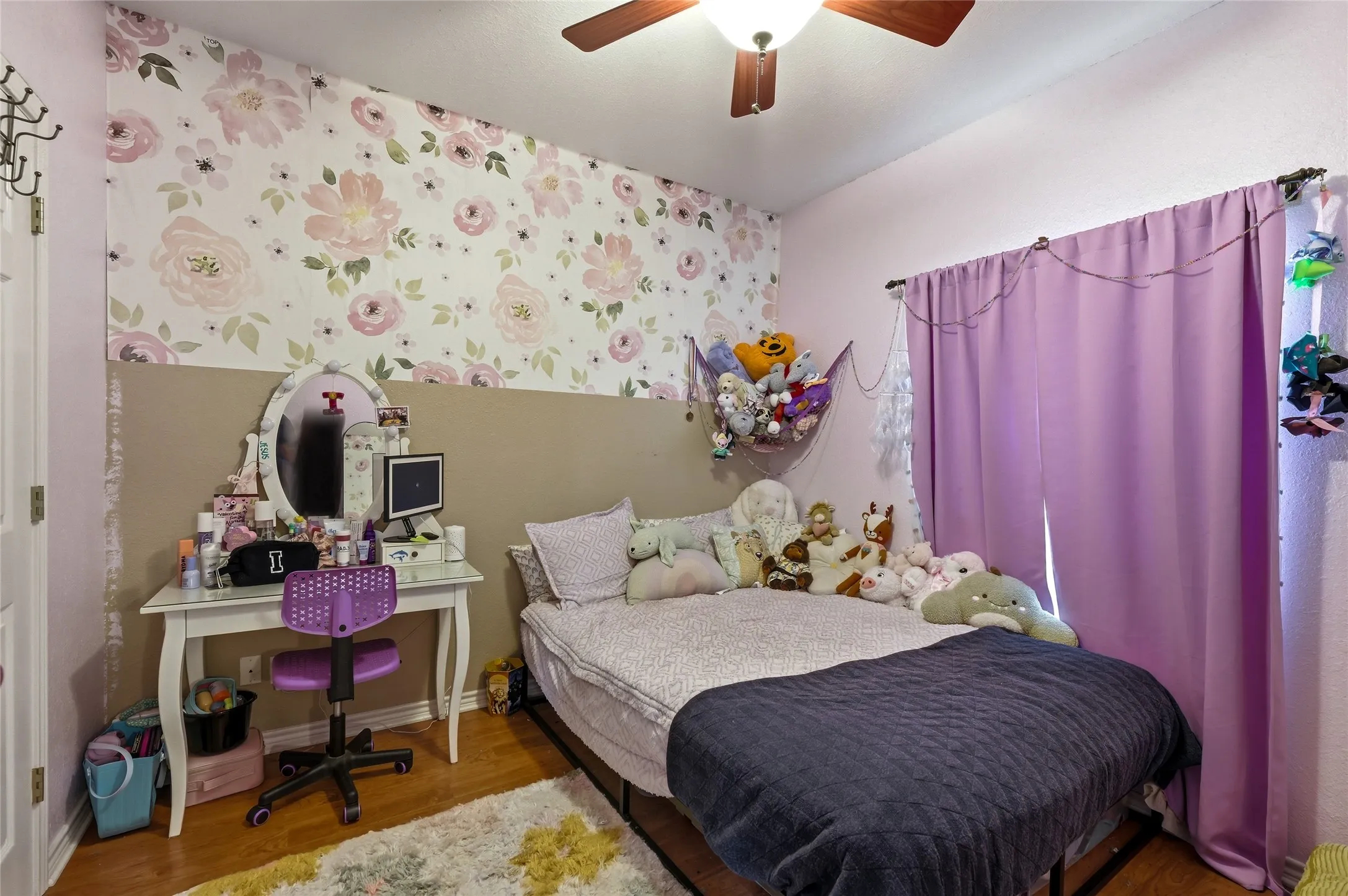 Interior space featuring a floral accent wall, wood-finish flooring, ceiling fan, window with curtains, and a vanity table with an oval mirror