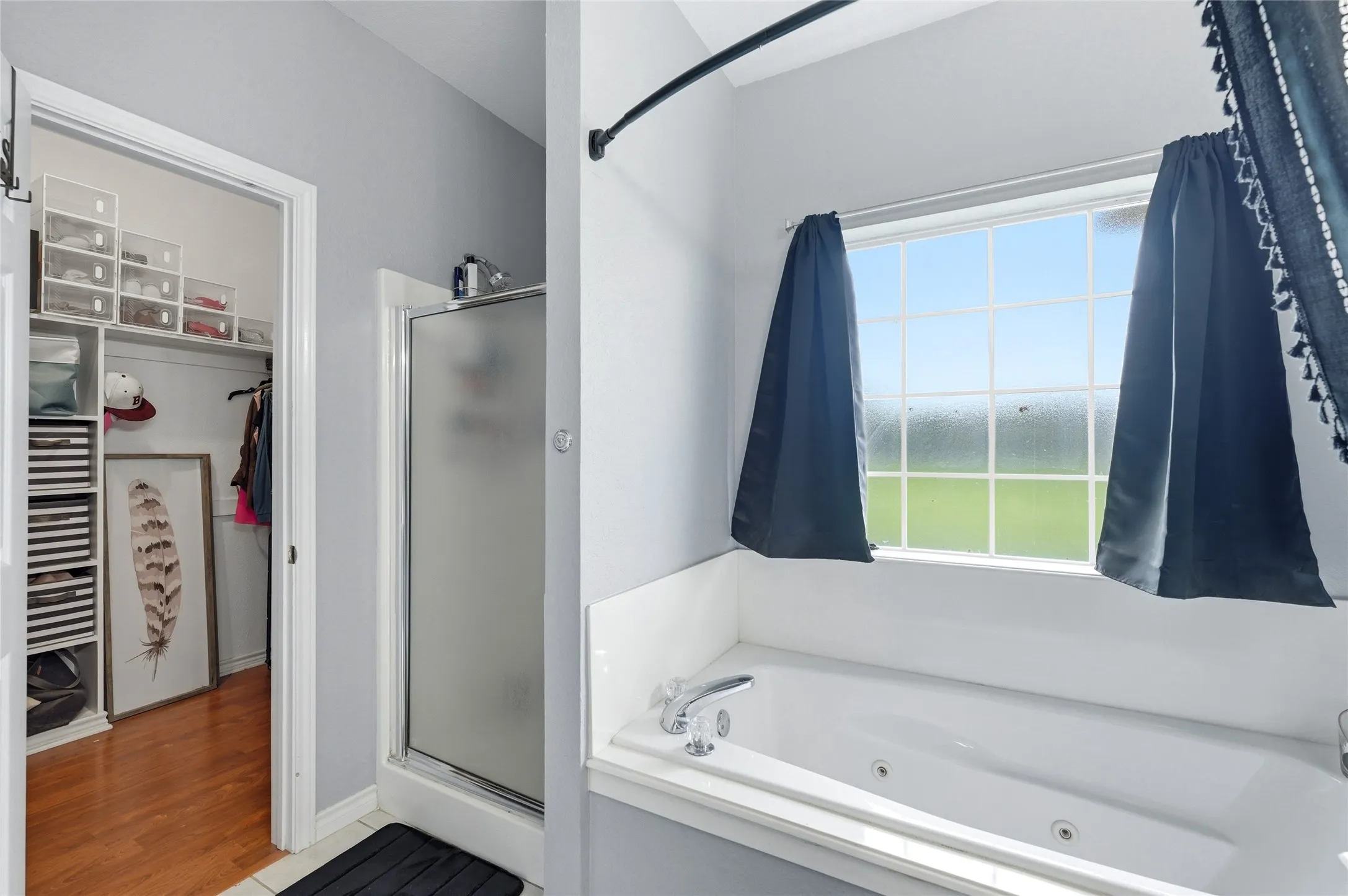 Soaking tub with a white tile surround and large window, accompanied by a glass-enclosed shower and a closet featuring wood-finish flooring and built-in shelving