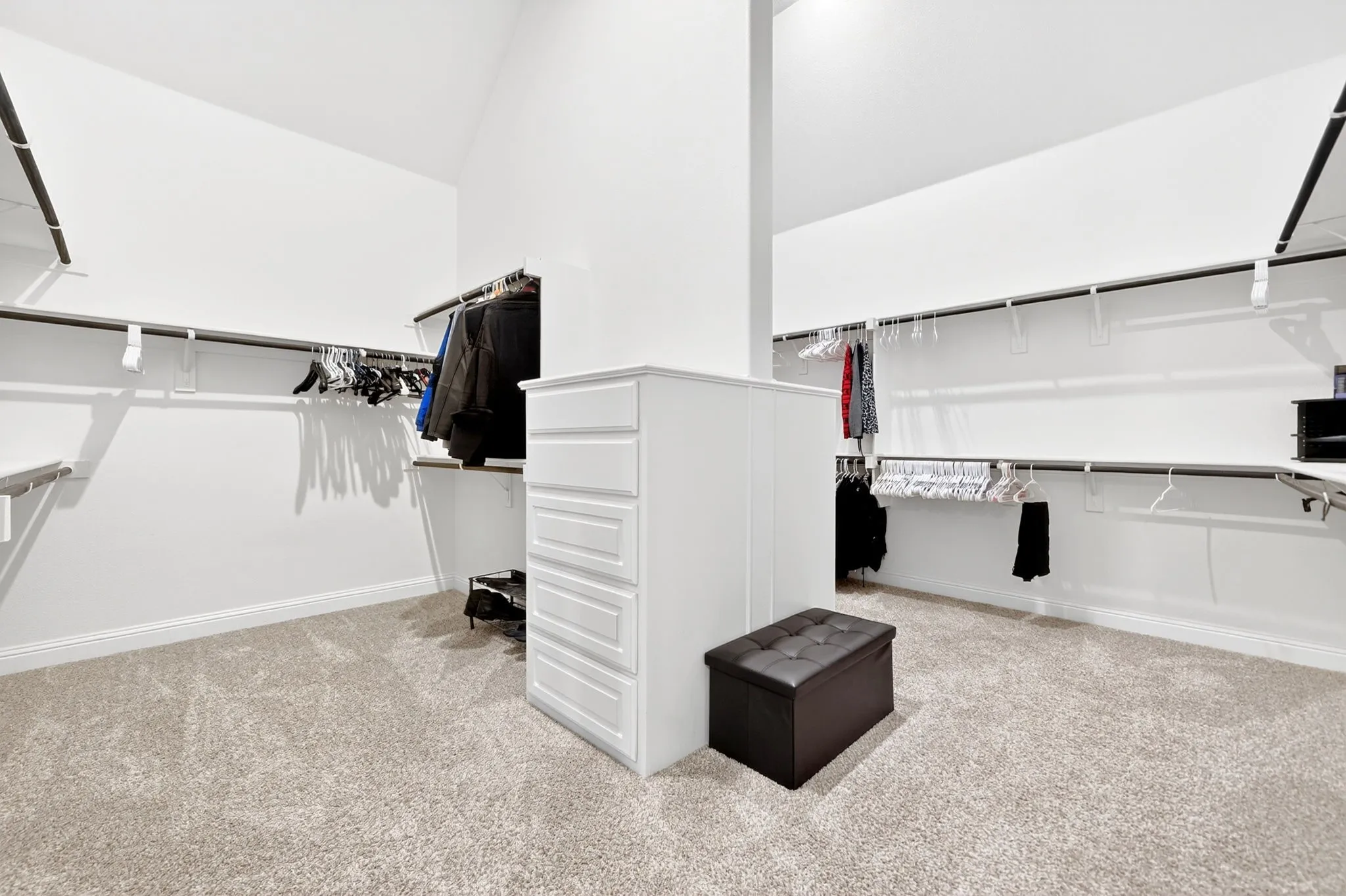 Spacious walk-in closet featuring extensive built-in shelving, multiple hanging rods, a central dresser island with drawers, and light-colored carpeting