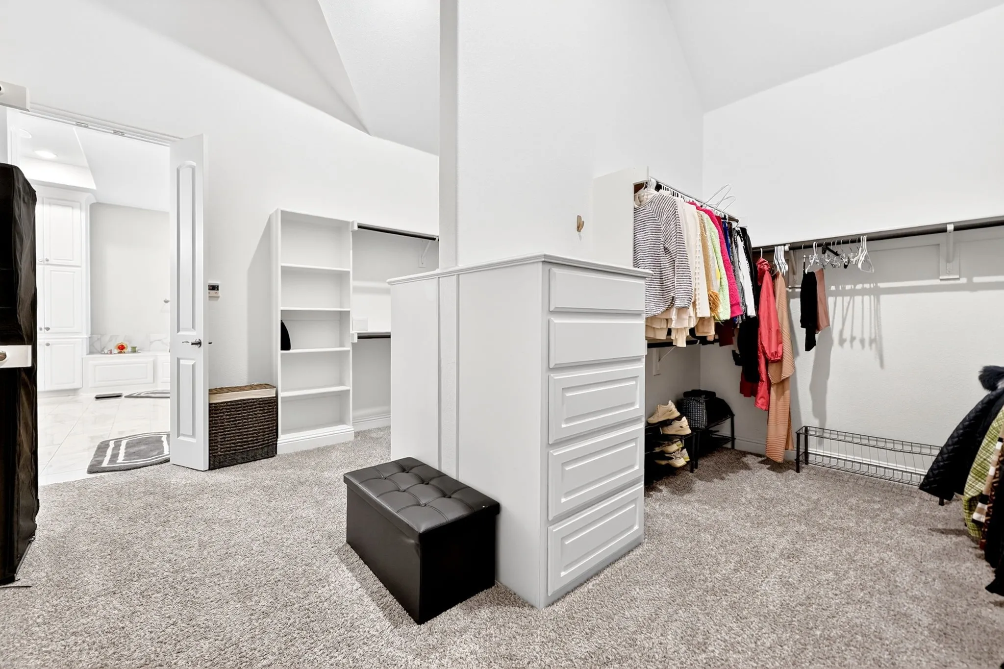 Carpeted walk-in closet featuring white built-in shelving, hanging rods, a central dresser, and a door leading to a tiled bathroom