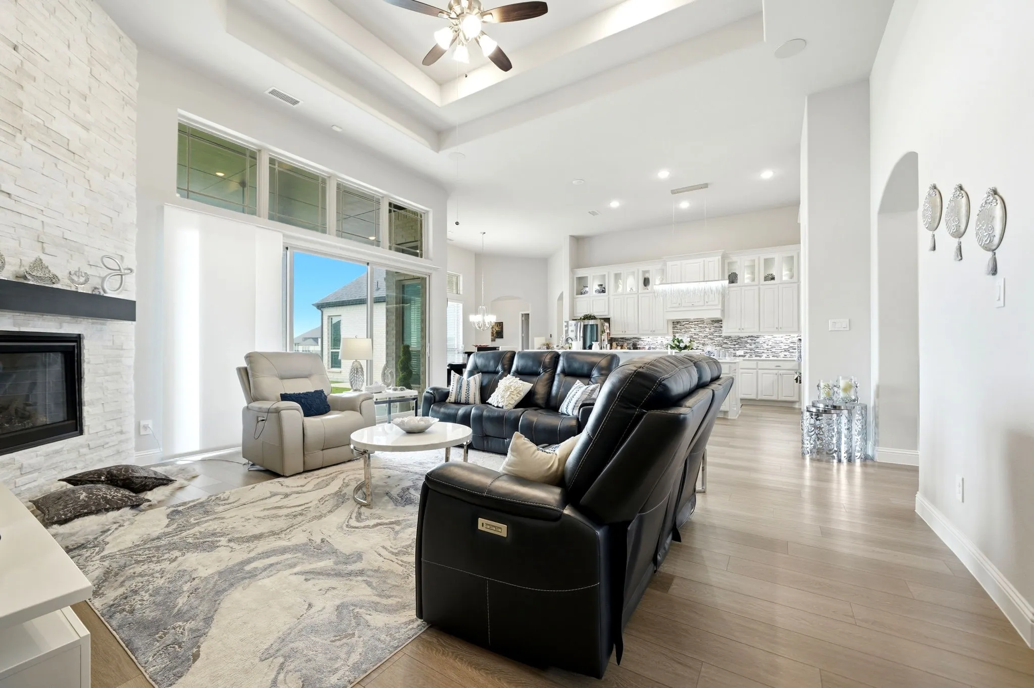 Open-concept living area featuring a stone-clad fireplace with dark mantel, wood-finish flooring, and a tray ceiling with a ceiling fan
