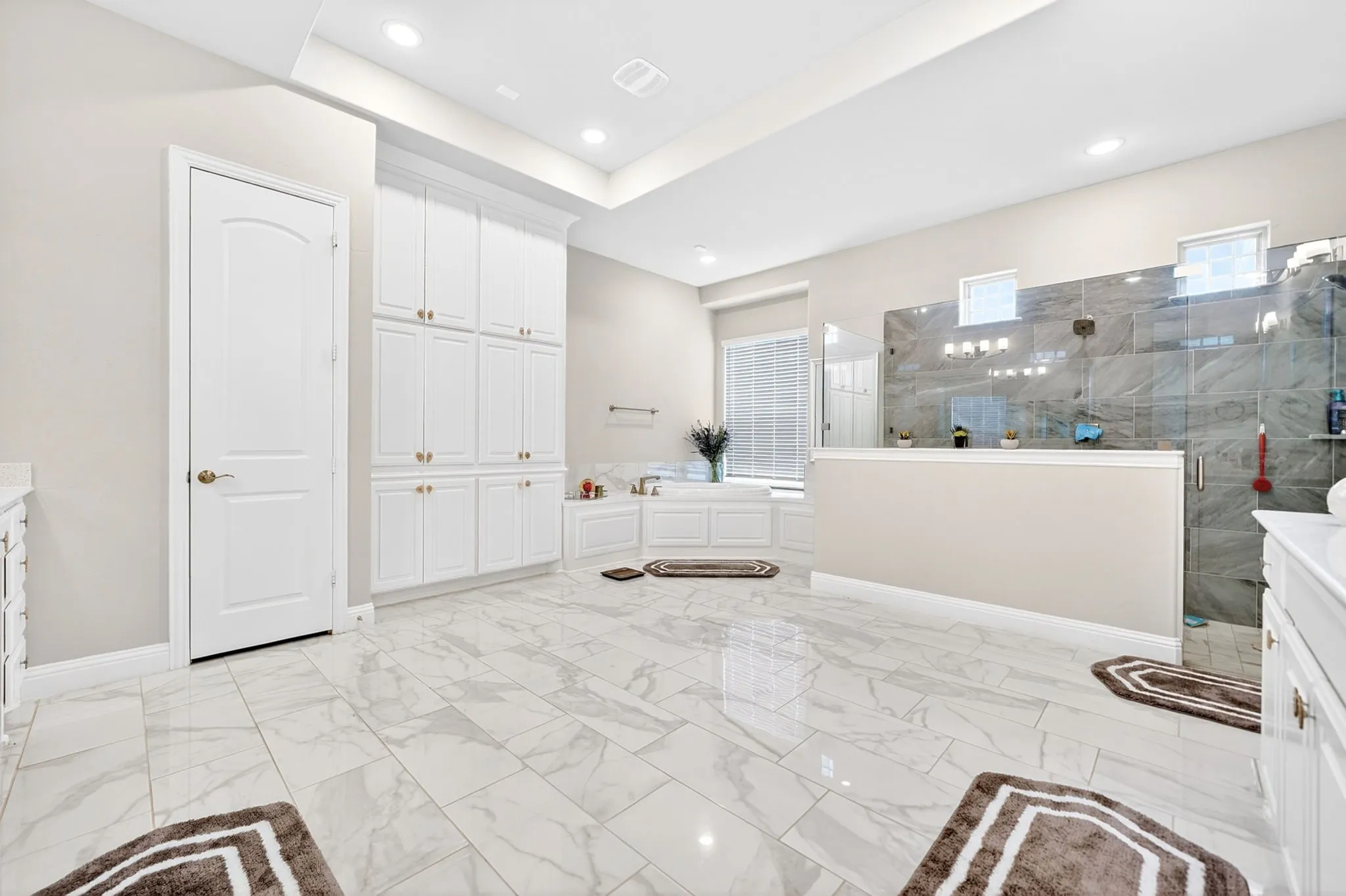 Spacious bathroom featuring large format marble-look tile flooring, extensive white built-in cabinetry, a walk-in shower with glass enclosure and gray tile surround, recessed lighting, and a garden tub with window