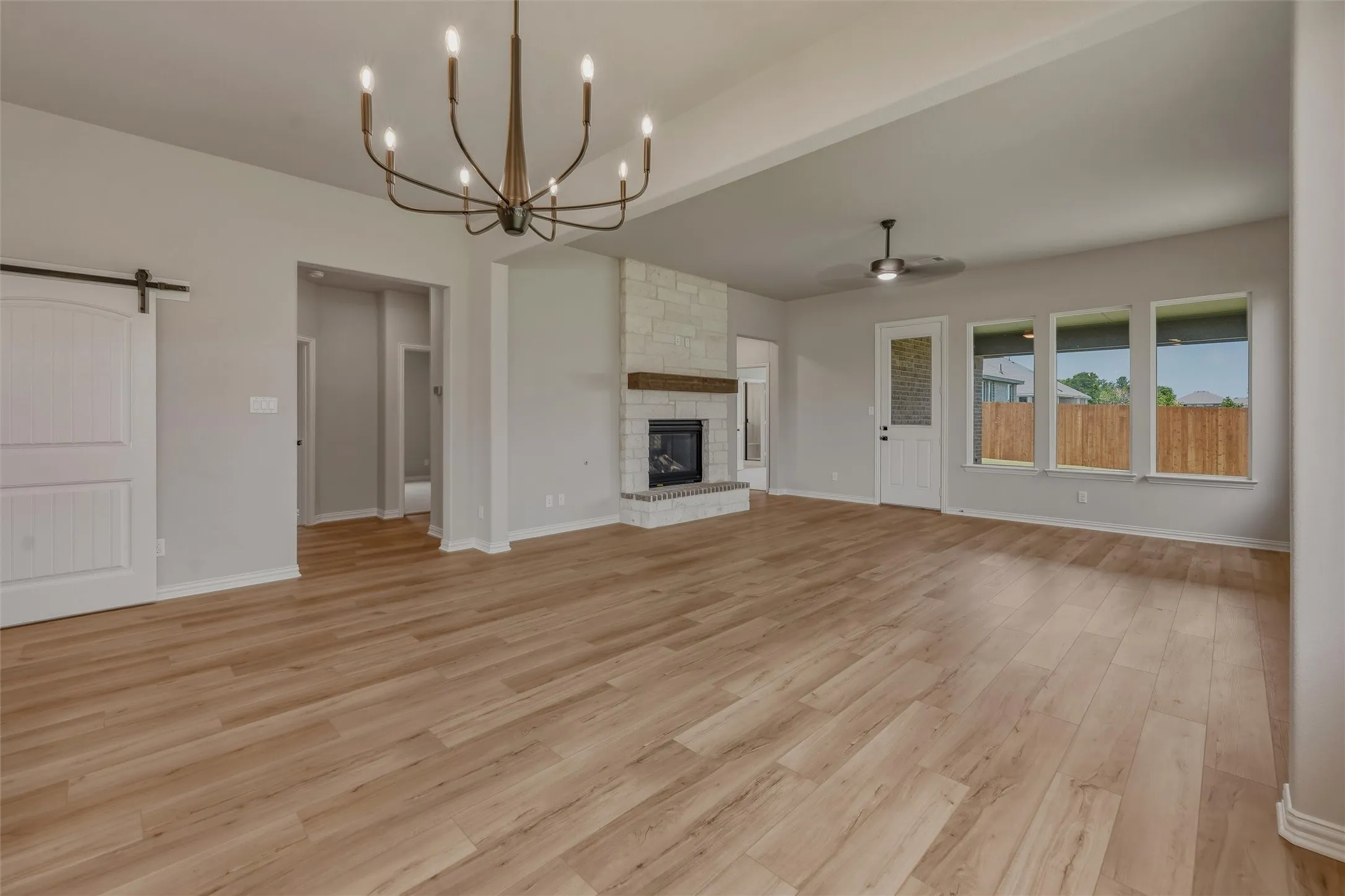 Spacious living area featuring wood-finish flooring, a stone-clad fireplace with a wood mantel, a modern chandelier, a ceiling fan, and multiple large windows