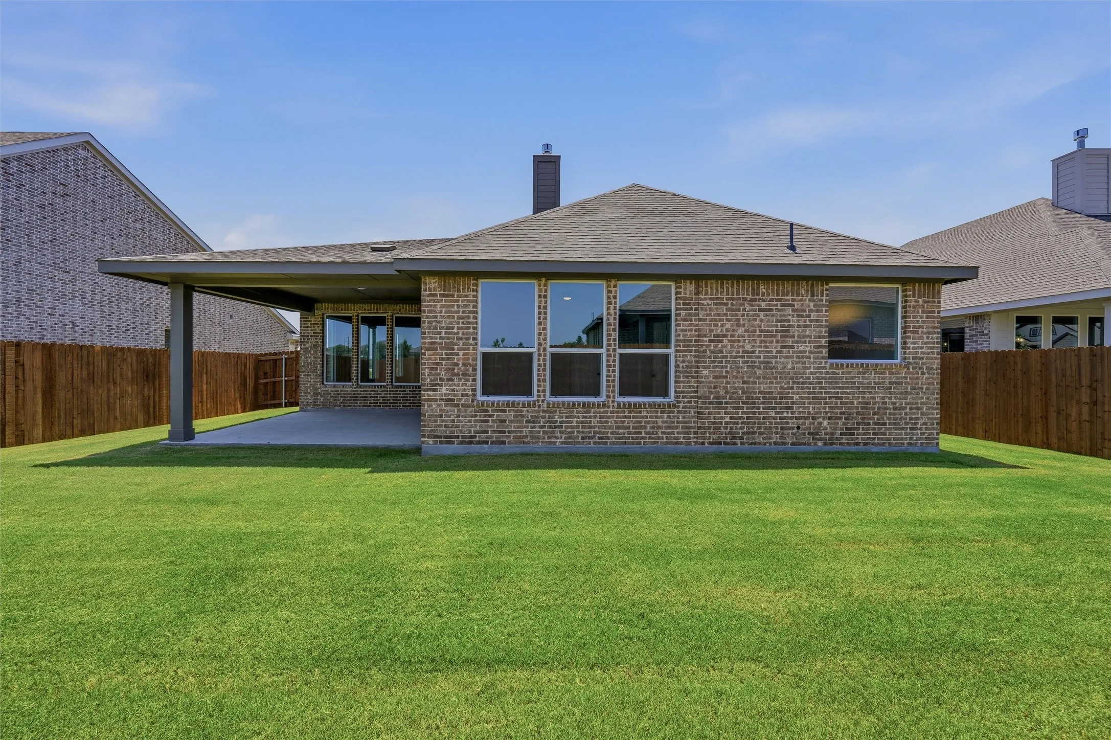 Expansive green lawn with a covered patio, brick exterior, dark-framed windows, and a privacy fence