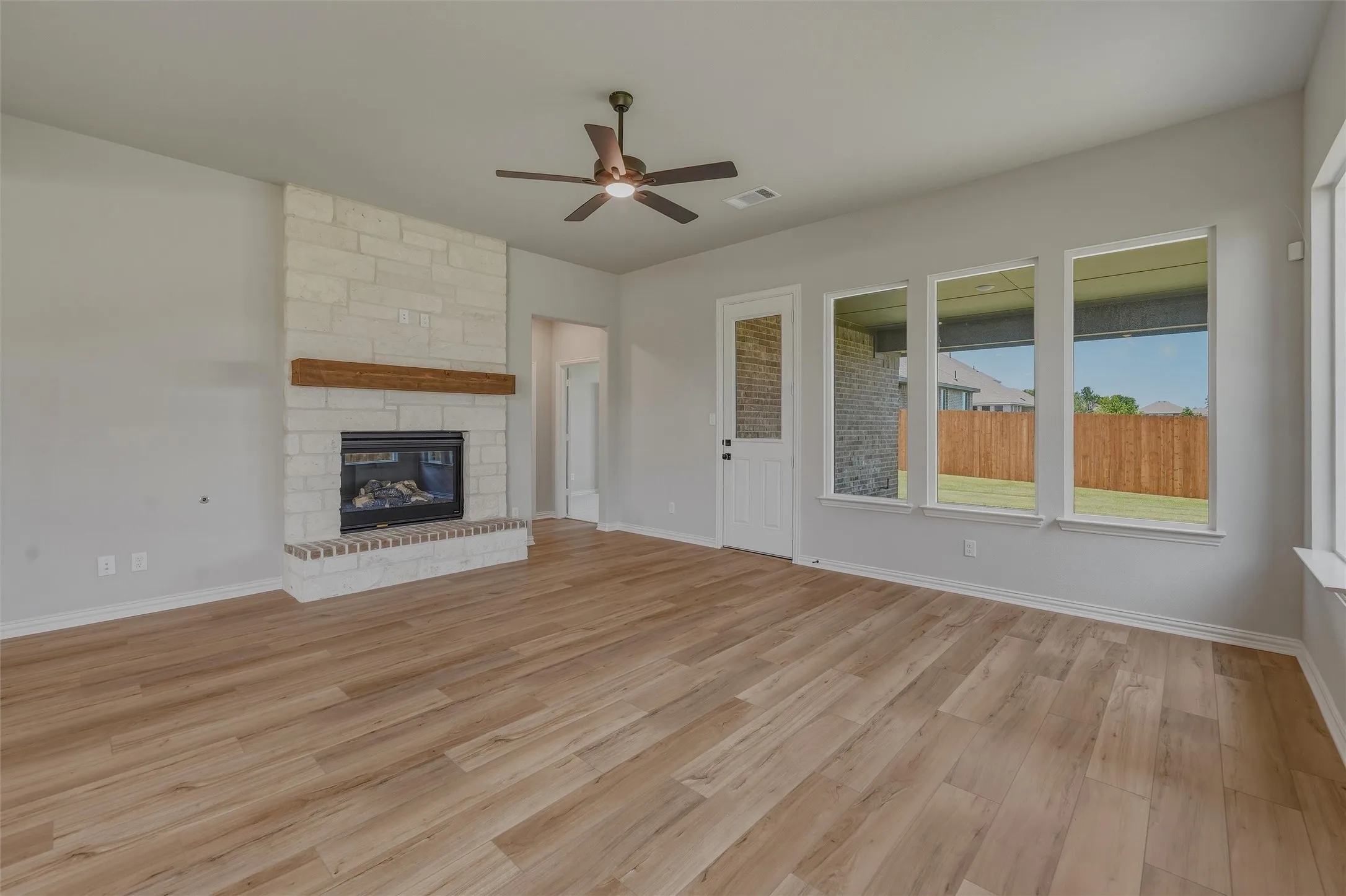 Spacious living area featuring light-tone wood-finish flooring, a stone-clad fireplace with a wood mantle, and a ceiling fan