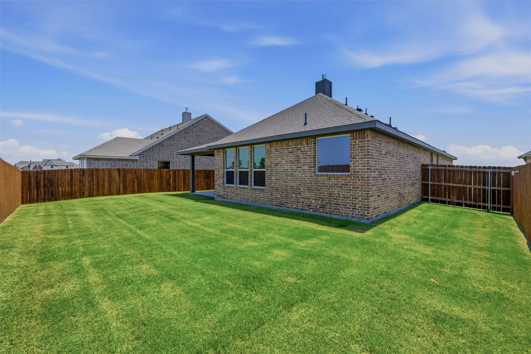 Expansive backyard featuring a manicured lawn, full privacy fencing, brick exterior, covered patio, and multiple windows