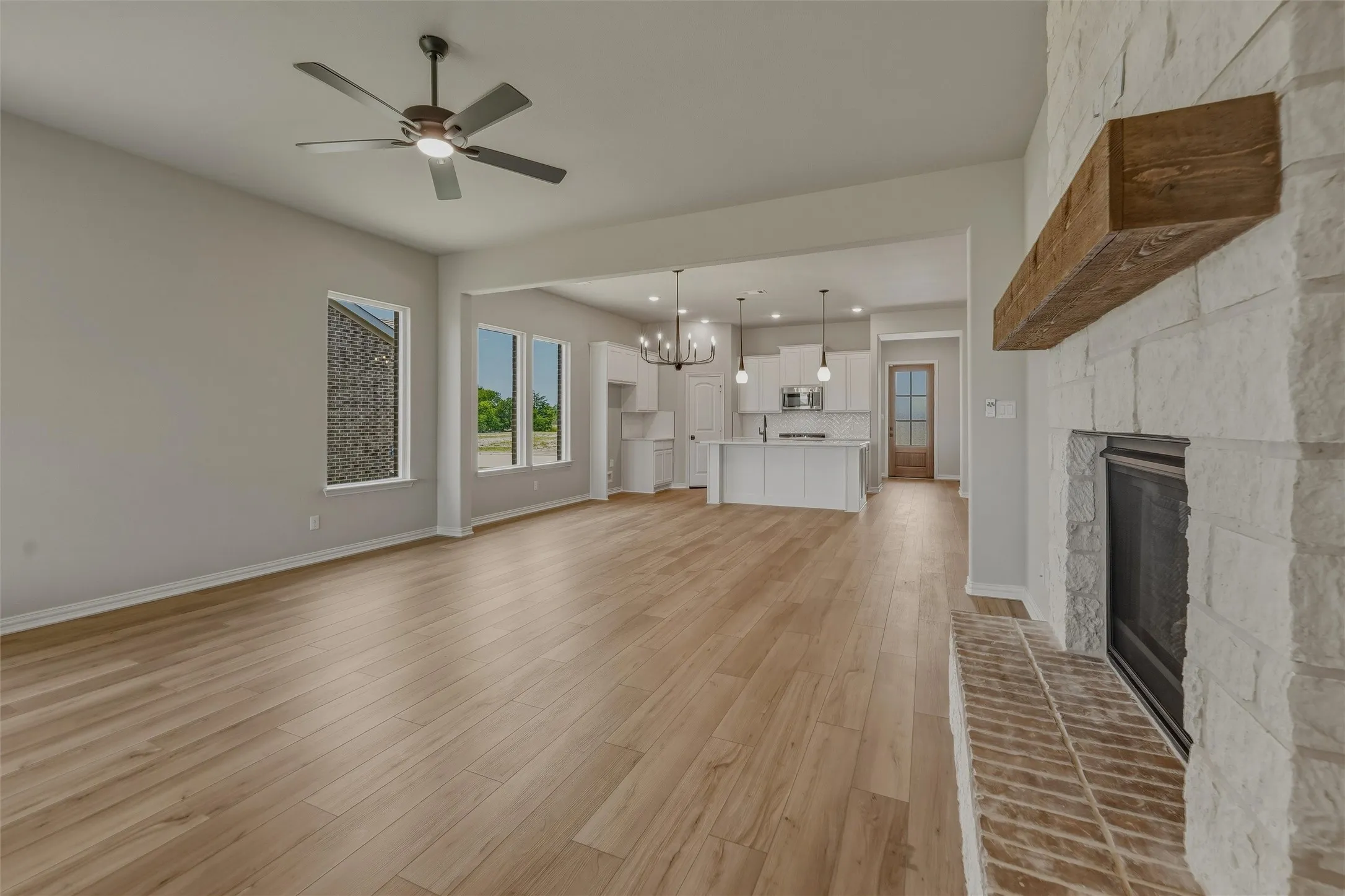 Open-concept living area featuring light wood-finish flooring, a stone-faced fireplace with a substantial wood mantel, a ceiling fan, and multiple large windows