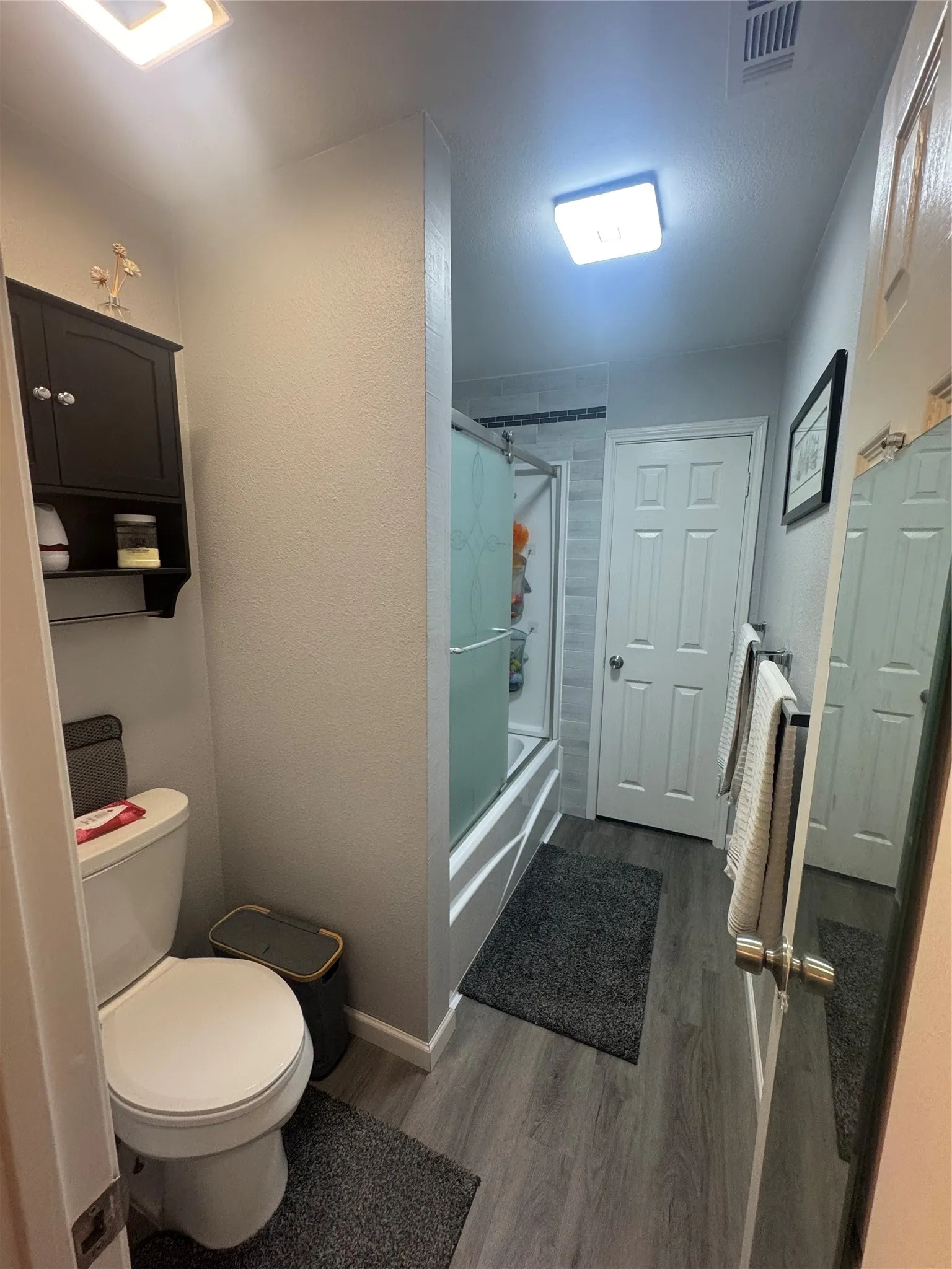 Bathroom featuring wood-finish flooring, a white toilet, a shower with sliding glass doors, and a dark wood-finish wall cabinet