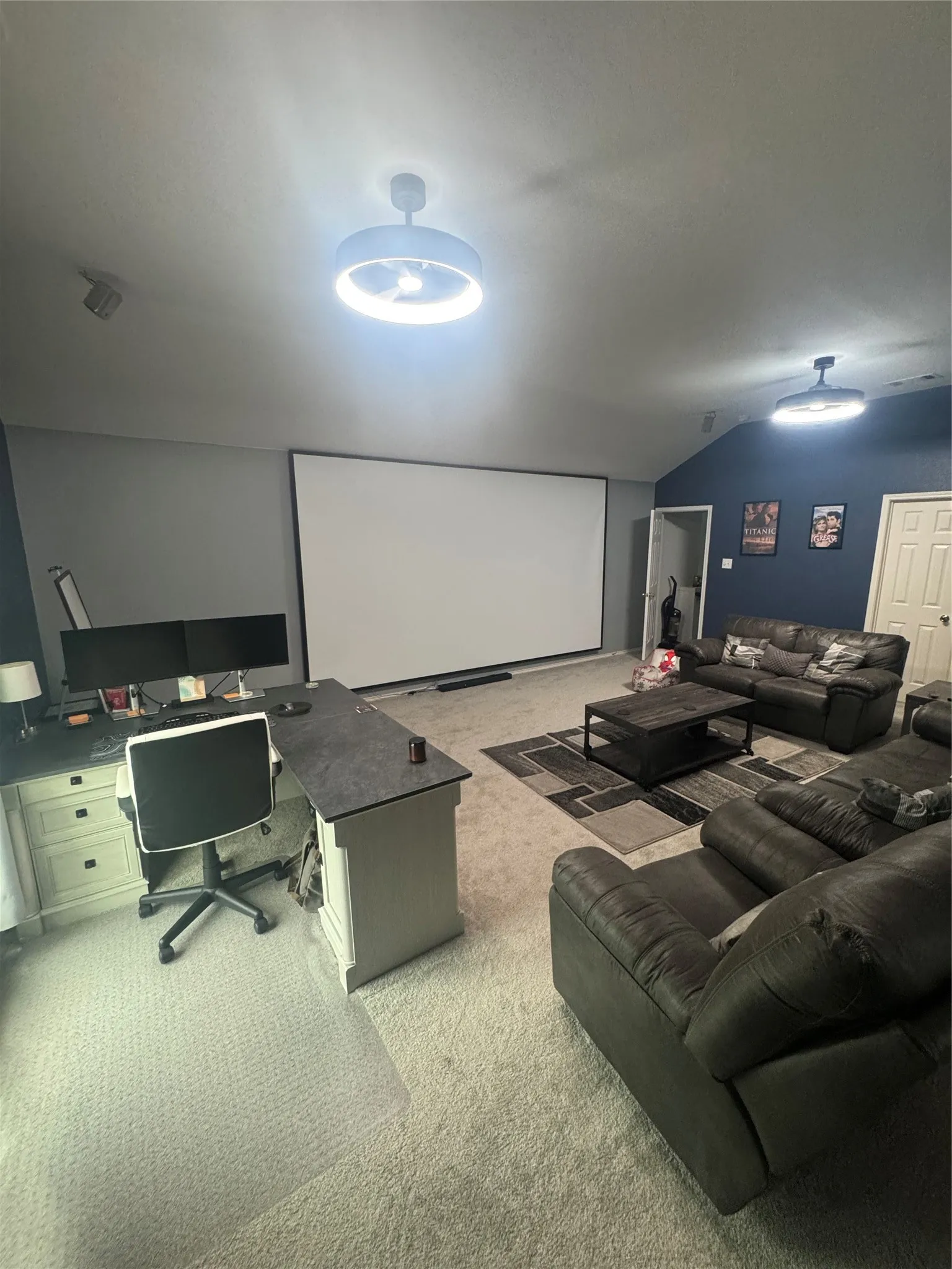 Spacious recreation room featuring a large projection screen, built-in desk with storage, two ceiling-mounted light fixtures, and sloped ceilings