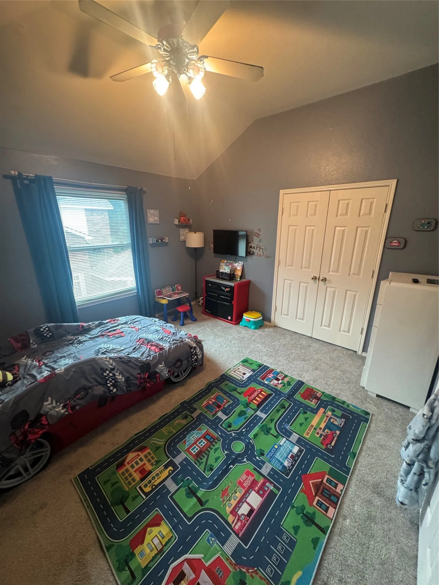 Carpeted room with a vaulted ceiling, a flush-mount ceiling fan with light fixture, a double-pane window with blue curtains, and a double-door closet