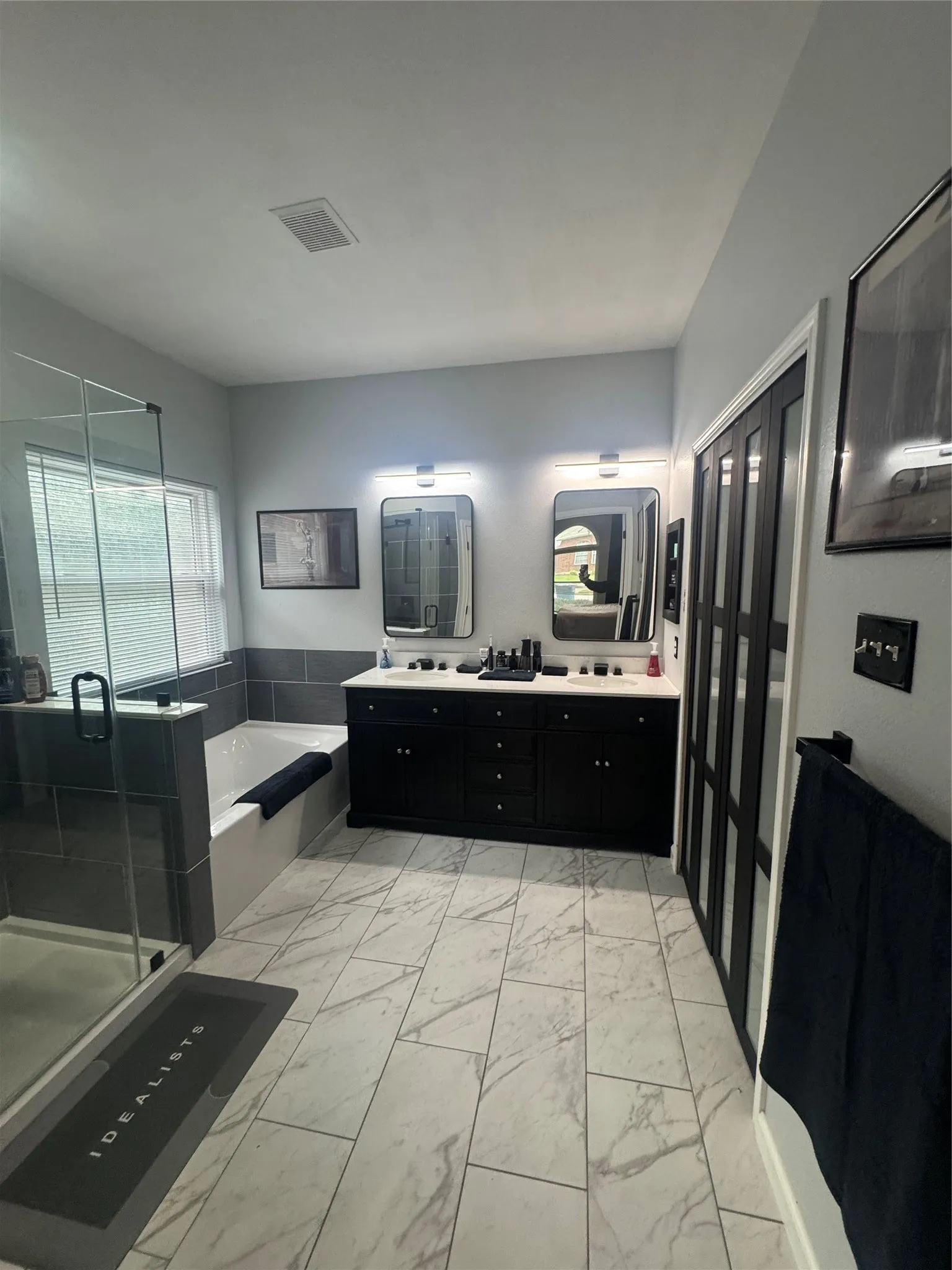 Spacious bathroom featuring marble-look tile flooring, a double vanity with ample storage, dual illuminated mirrors, a soaking tub with tiled surround, and a glass-enclosed shower