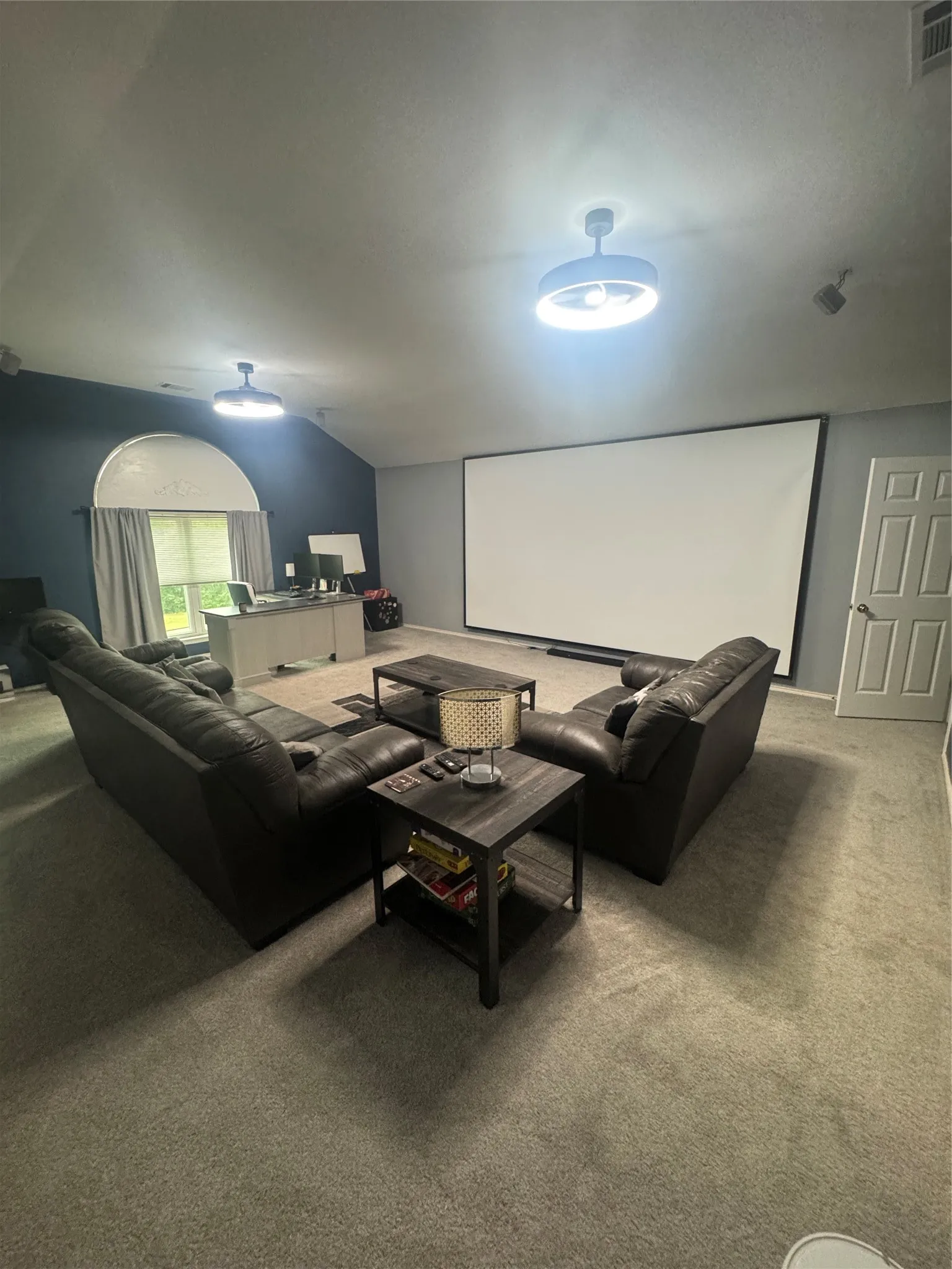 Spacious entertainment room featuring a large projection screen, neutral carpet flooring, and an arched window with drapes