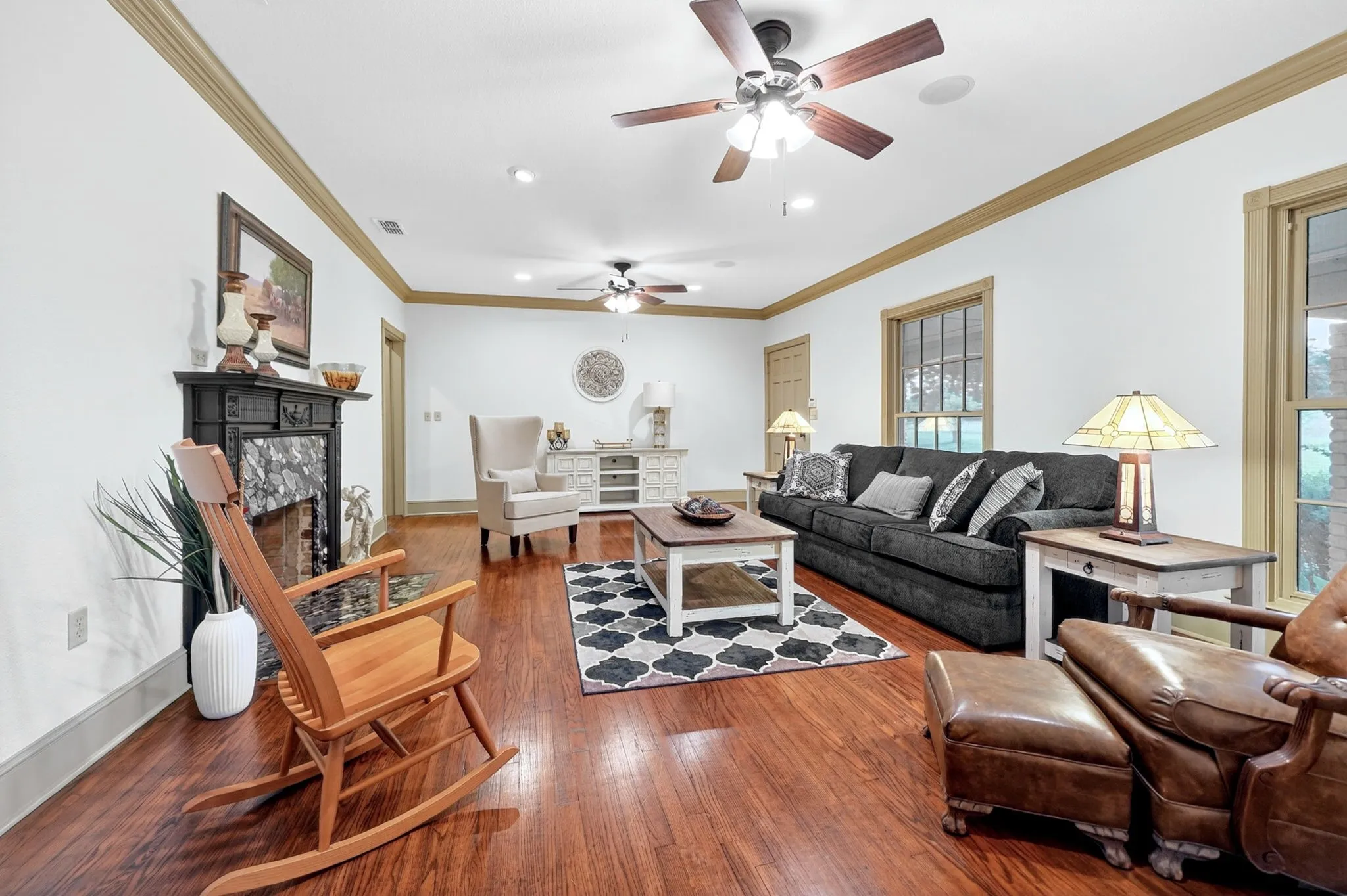 Spacious living area featuring polished wood-finish flooring, a decorative fireplace with a dark mantel and stone surround, and multiple ceiling fans