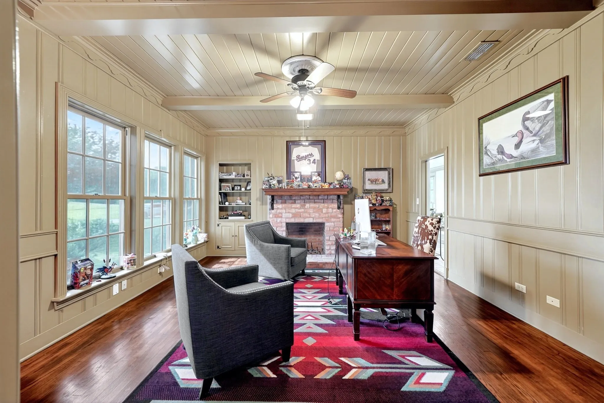Room featuring wood-finish flooring, a brick fireplace, built-in shelving, paneled walls, and multiple grid-pattern windows