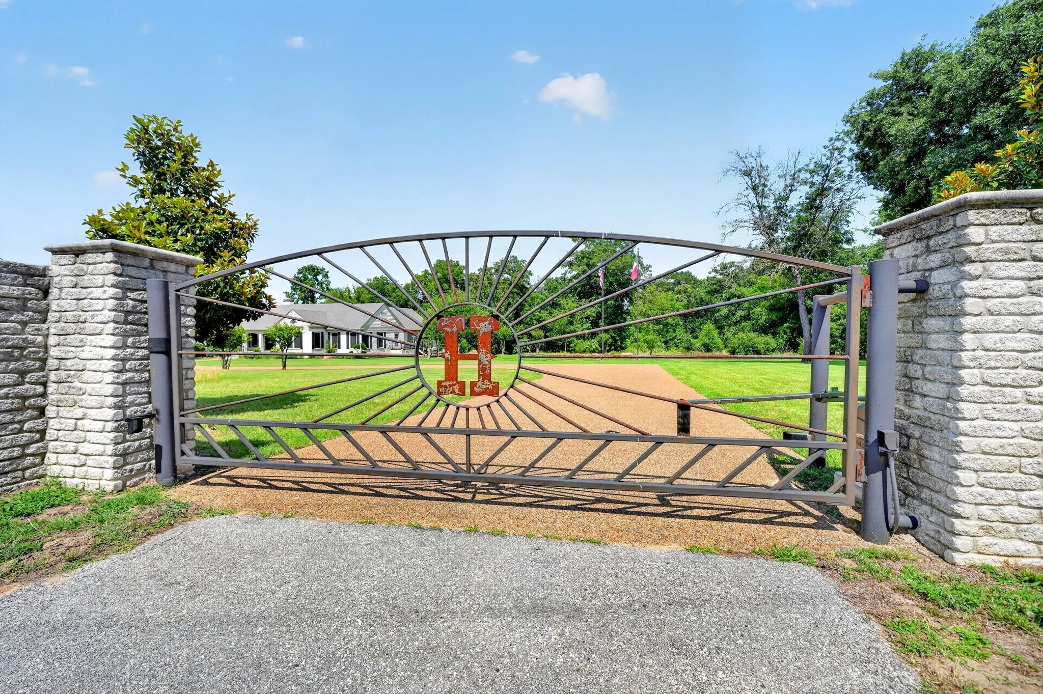 Automated metal gate with stone-clad pillars, gravel driveway, and expansive manicured lawn