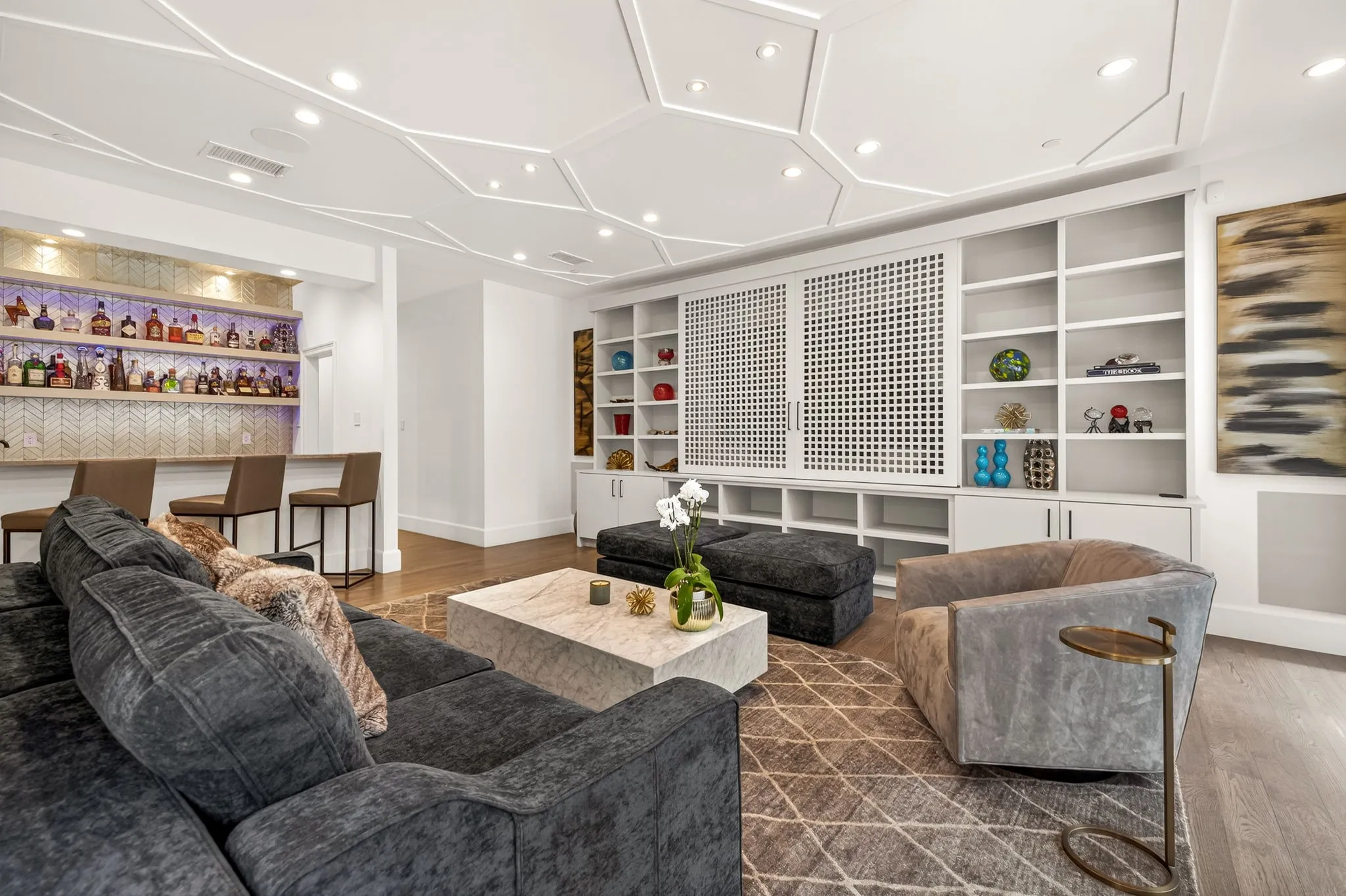 Entertainment room featuring a built-in wet bar with herringbone tile backsplash, custom cabinetry with integrated shelving, geometric coffered ceiling with recessed lighting, and wood-finish flooring