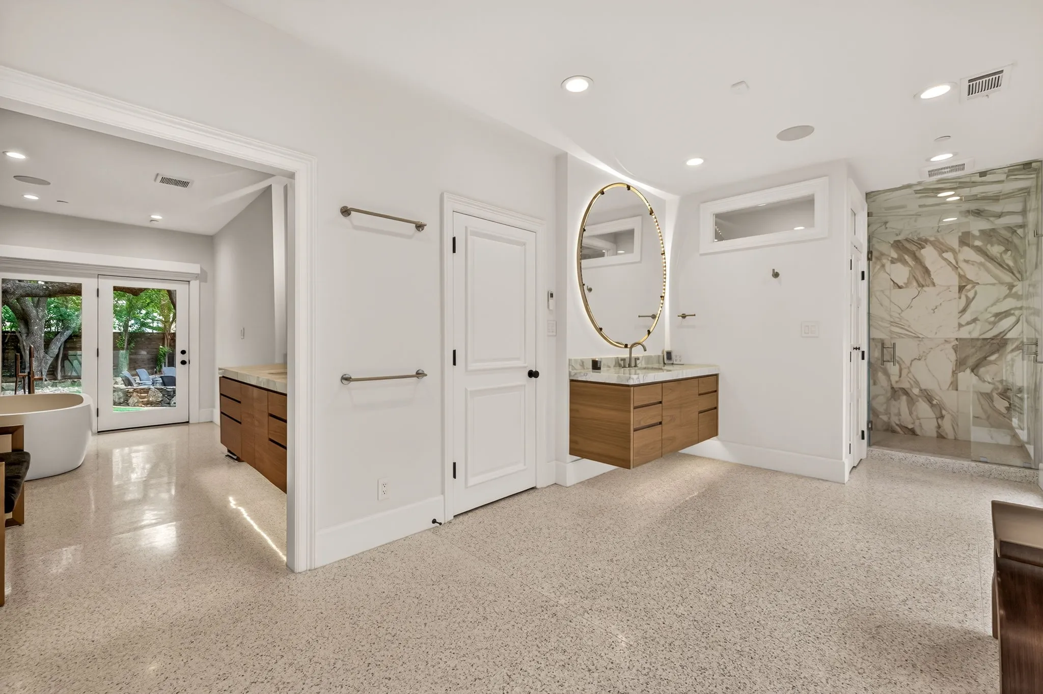 Spacious bathroom featuring light-toned speckled flooring, a floating wood-finish vanity with an oval illuminated mirror, and a walk-in shower with stone-patterned tile walls and a glass enclosure