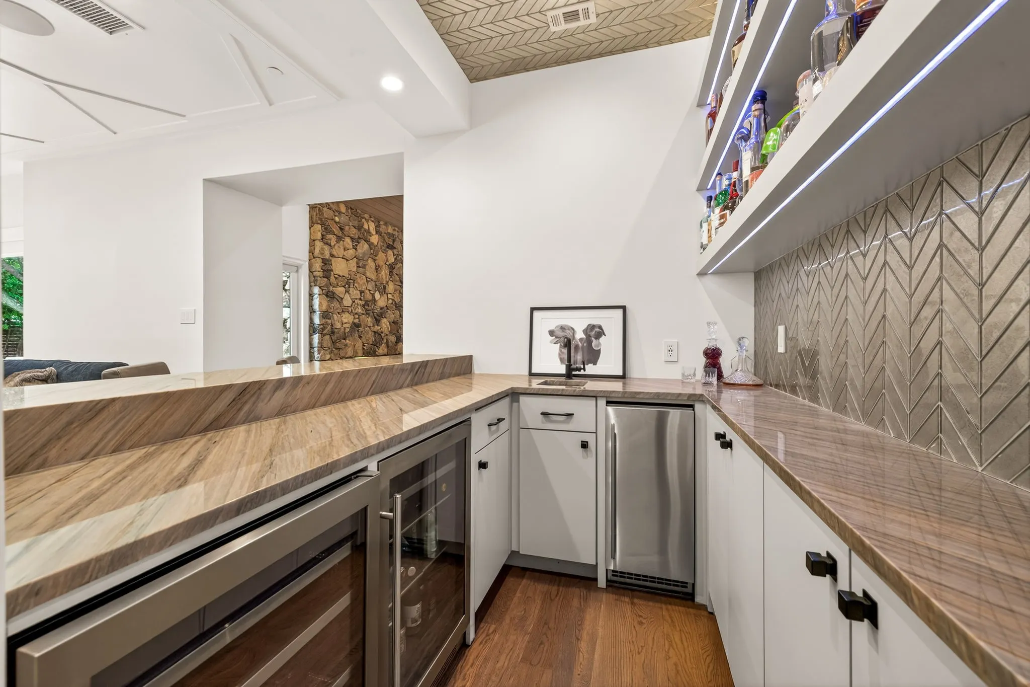 Custom bar area featuring stone countertops, built-in beverage refrigerator, wine cooler, herringbone tile backsplash, and integrated shelving with lighting
