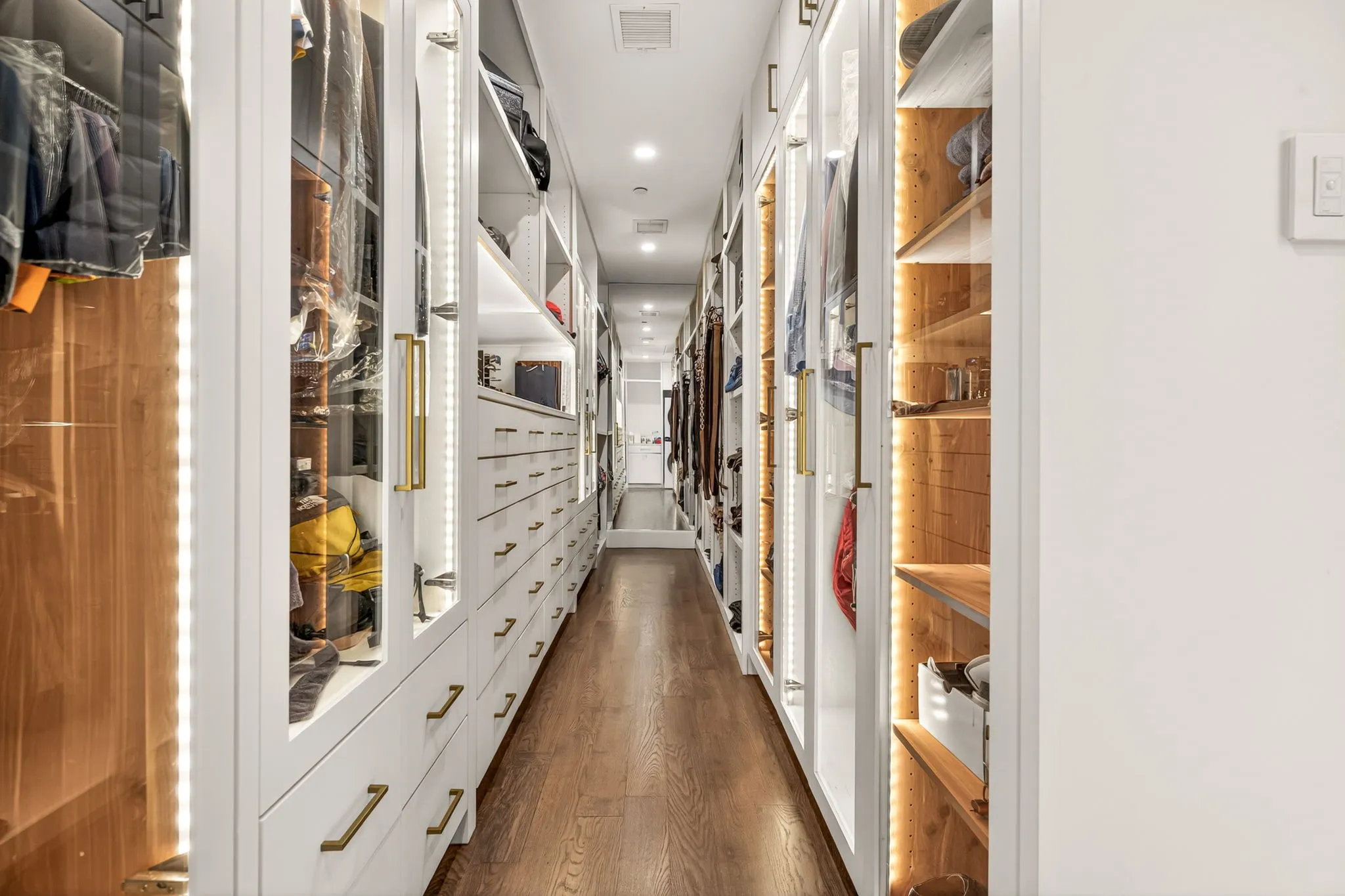 Customized closet system featuring glass-front doors, integrated lighting, wood-finish shelving, an array of drawers, and polished brass hardware