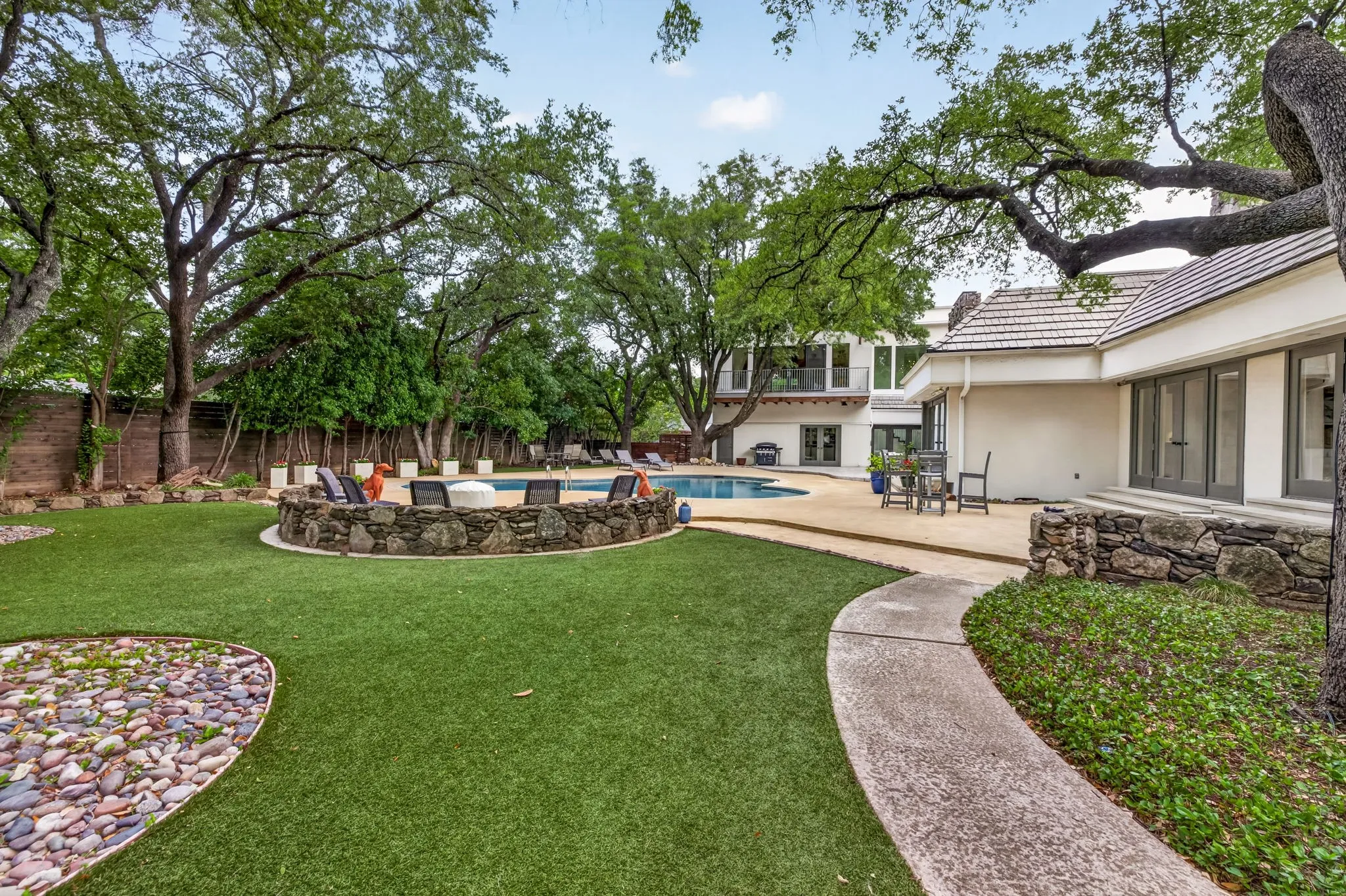 Expansive backyard featuring artificial turf, a stone-lined fire pit, a swimming pool, and mature trees