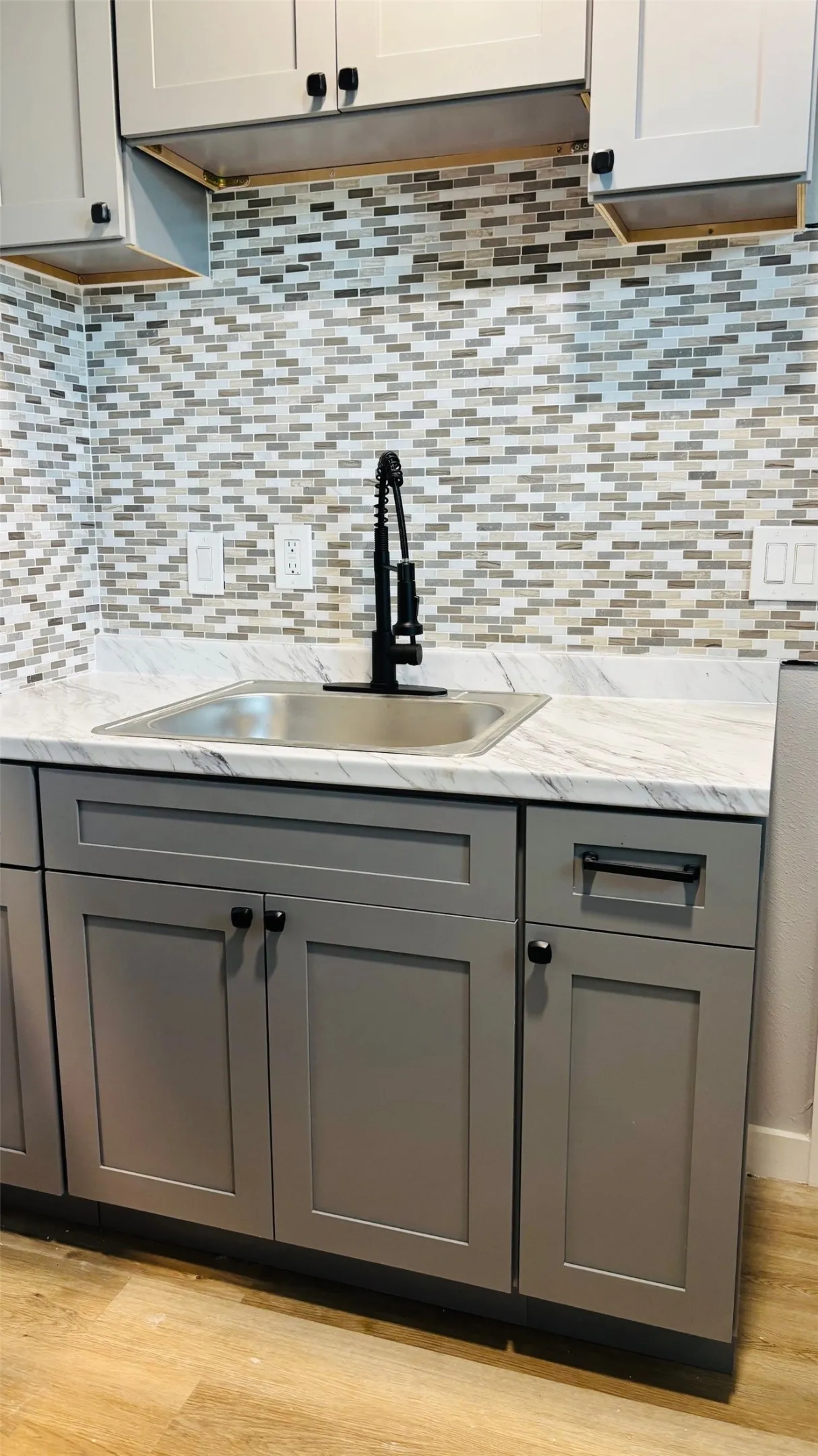 Kitchen sink area featuring gray lower cabinetry, light upper cabinetry, a stainless steel sink, a black gooseneck faucet, and a mosaic tile backsplash