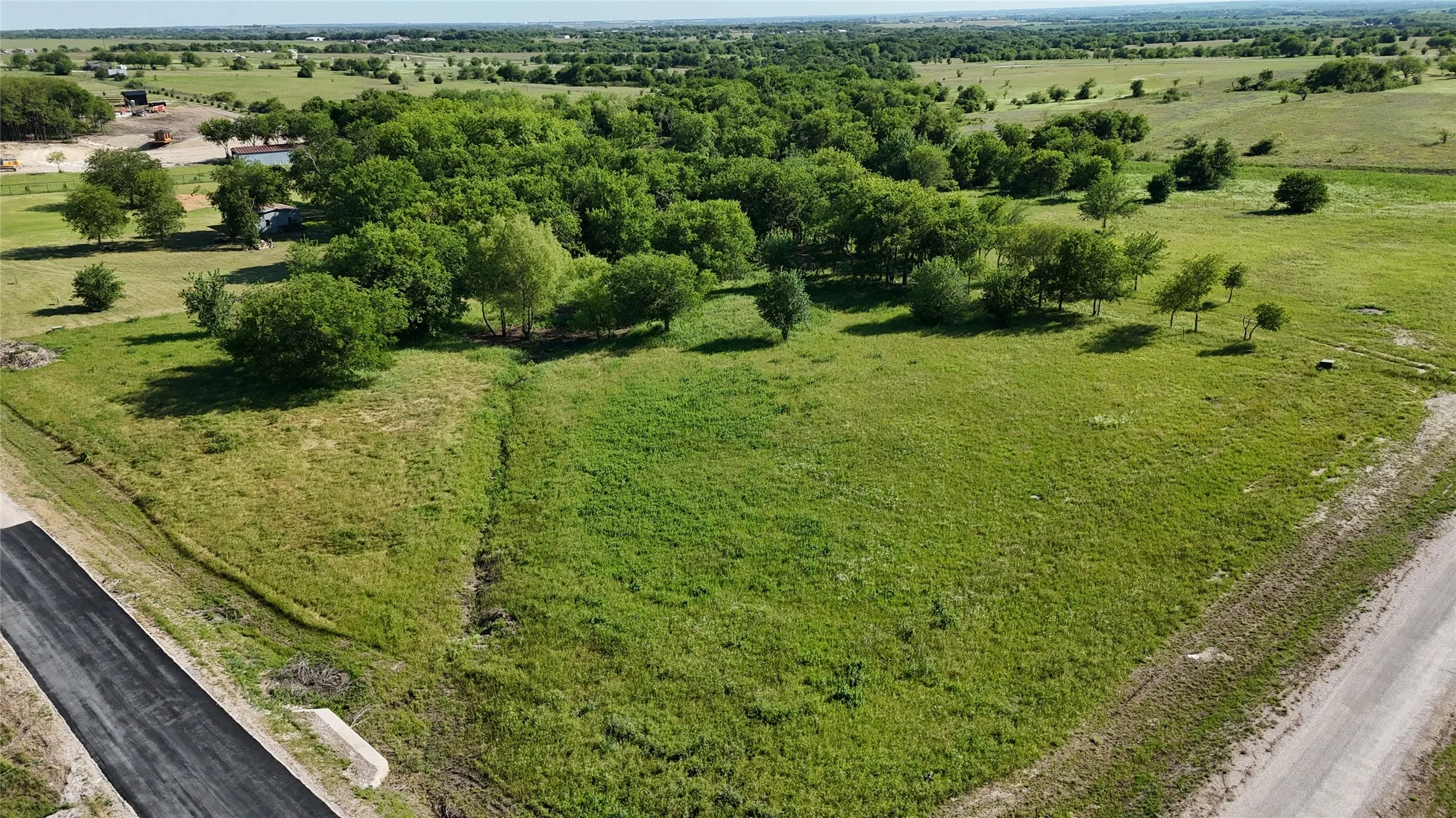 Expansive green acreage featuring a treeline, paved road frontage, and a gravel road border