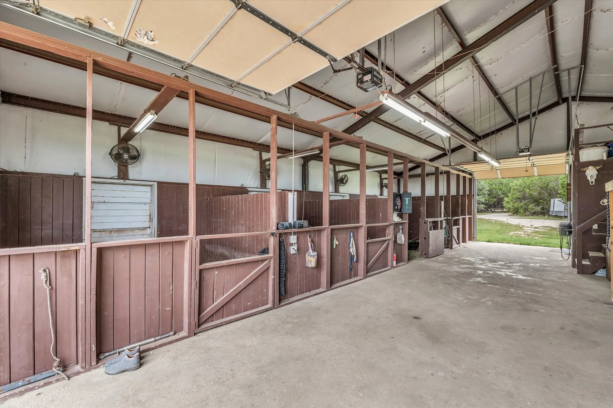 Spacious barn interior featuring multiple stalls with wood paneling, concrete flooring, exposed metal beam ceiling, and fluorescent overhead lighting